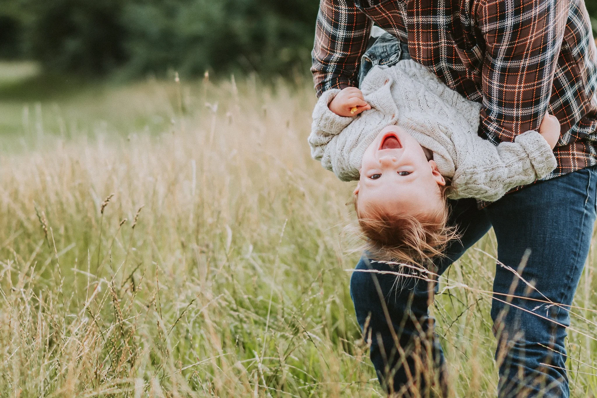 Child hanging upside down with a happy expression, held by an adult in a grassy outdoor area.