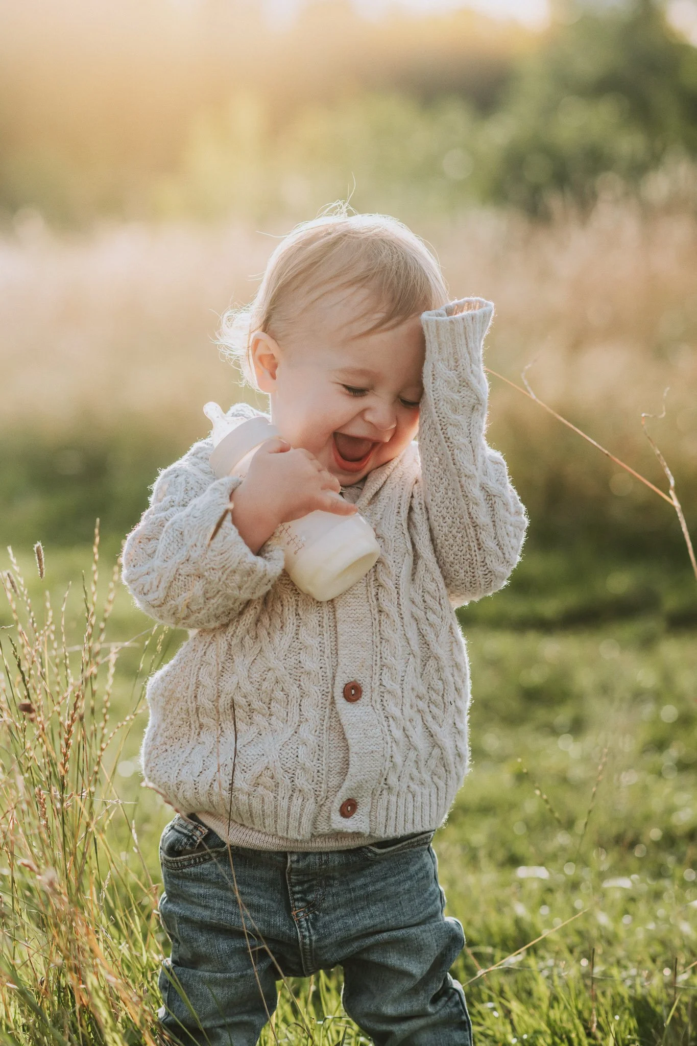 A young child holding a baby bottle, smiling, and wiping their forehead with one hand outdoors in a grassy field during sunset.