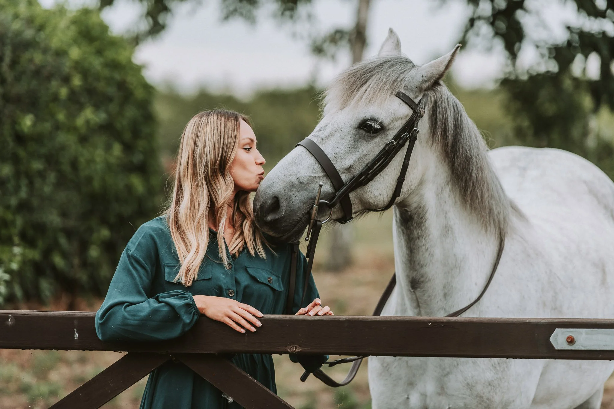 A woman with shoulder-length blonde hair wearing a dark teal jacket kisses a white horse with gray mane on the nose over a wooden fence, outdoors with trees in the background.