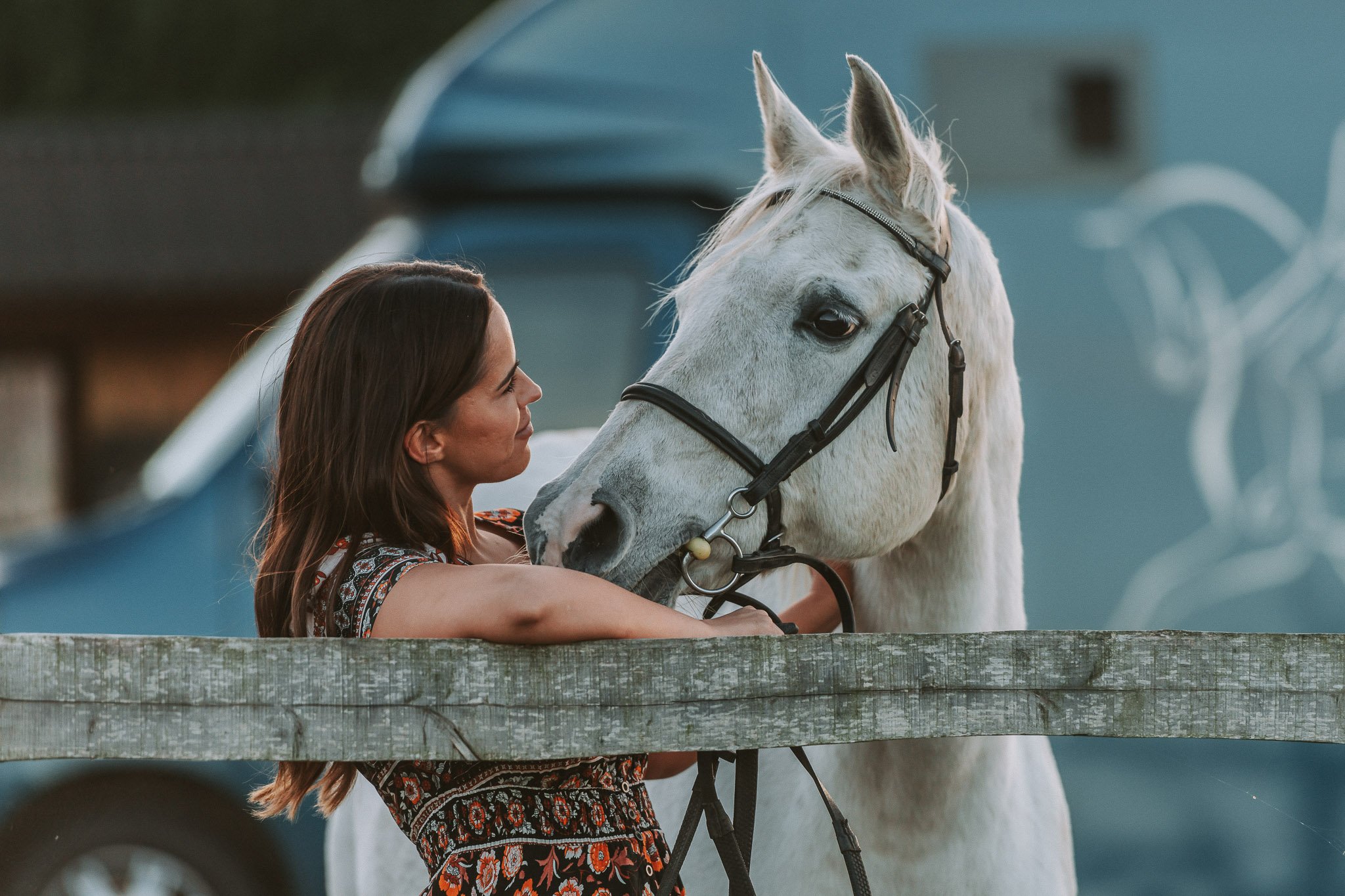 A woman with brown hair leaning on a wooden fence and smiling at a white horse with a black bridle.