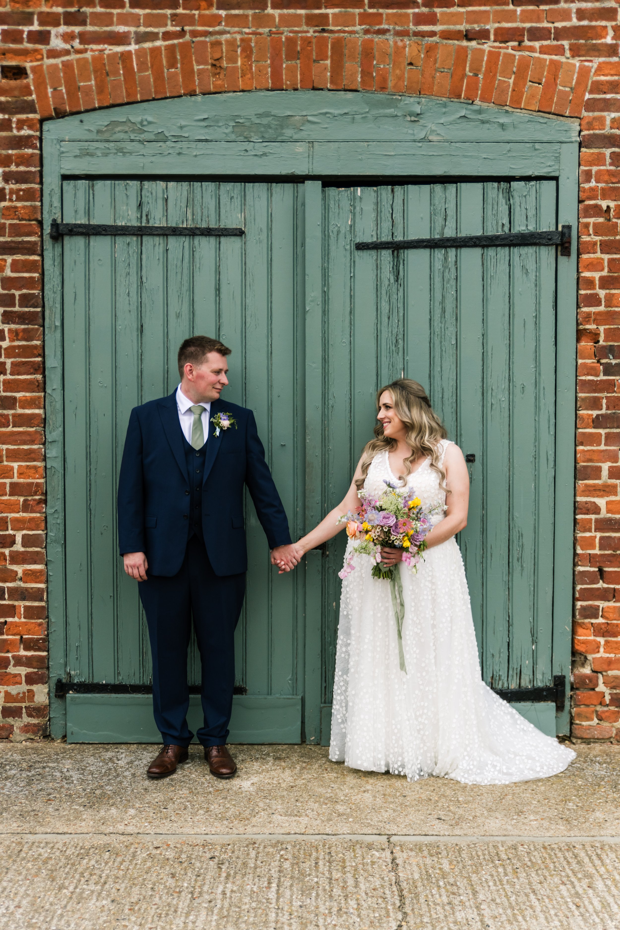 A bride and groom holding hands in front of green wooden doors, with brick walls on sides. The bride is in a white wedding dress holding a bouquet, and the groom is in a navy blue suit.