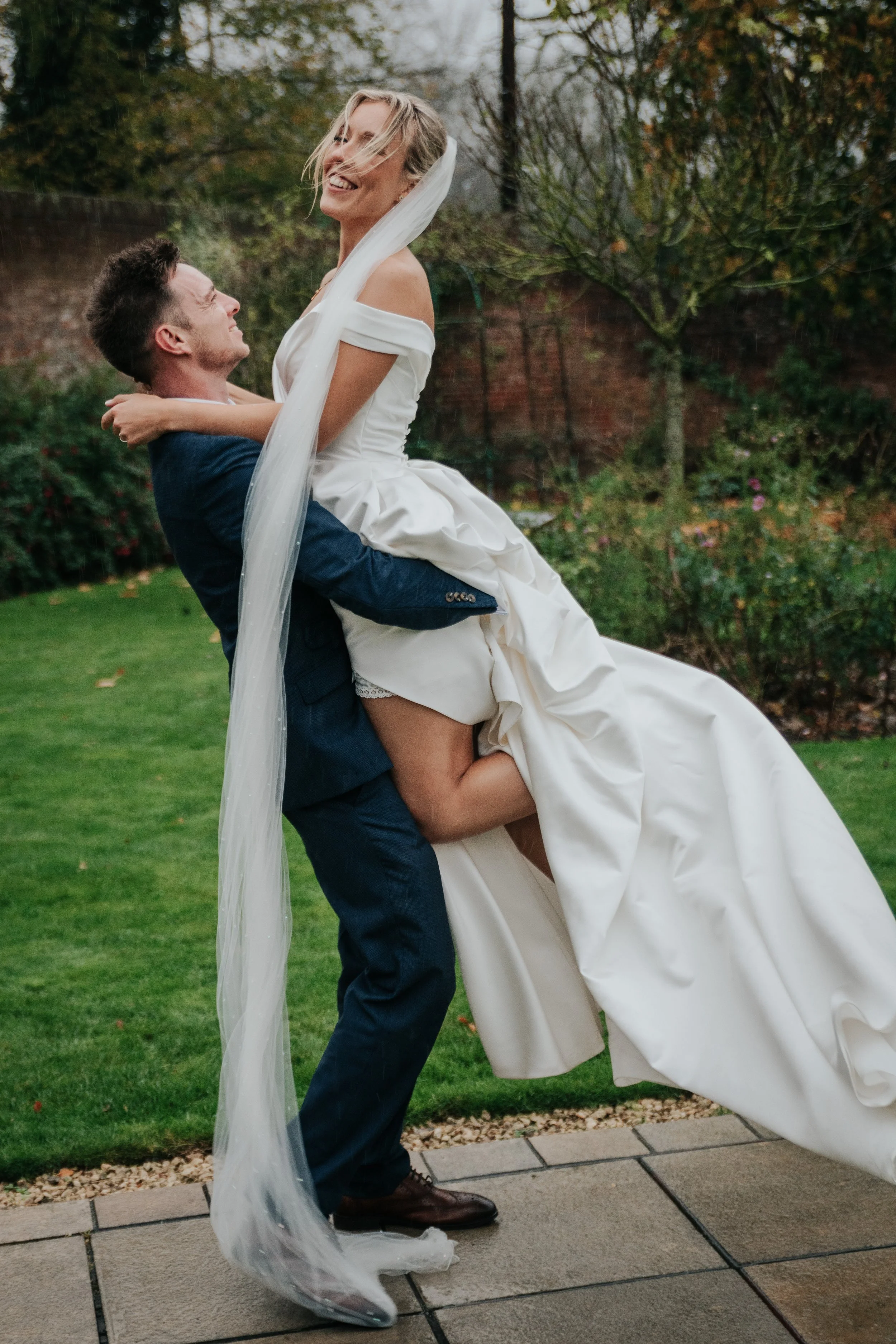 A man in a blue suit is lifting a woman in a white wedding dress in a garden with trees and a brick fence. The woman is smiling and wearing a veil.