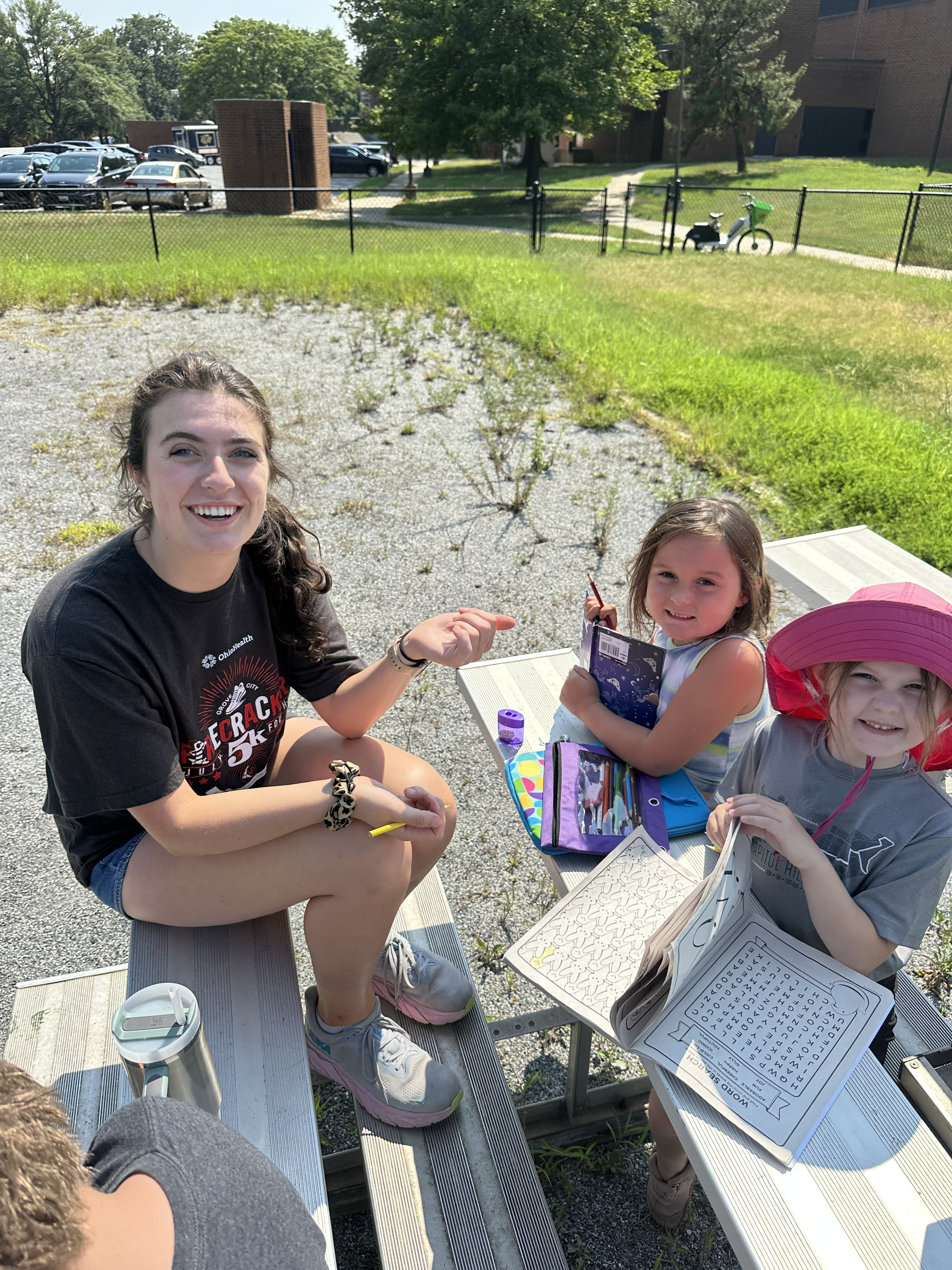 Smiling young woman and two young girls sitting at outdoor picnic tables with books and coloring materials on a bright, sunny day.