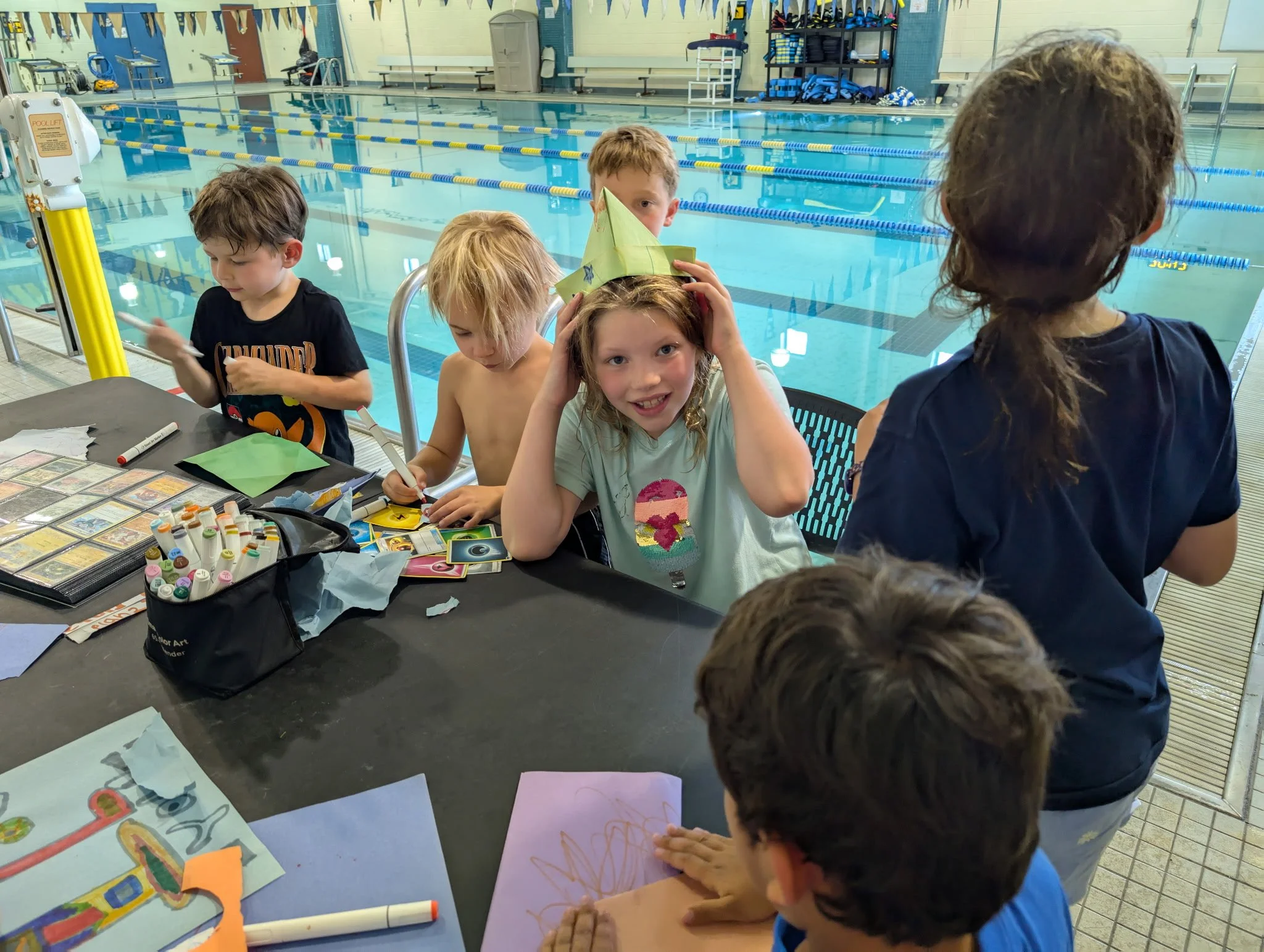 Group of children at a swimming pool side table participating in a crafting activity with various art supplies, some children drawing and one girl adjusting a paper hat.