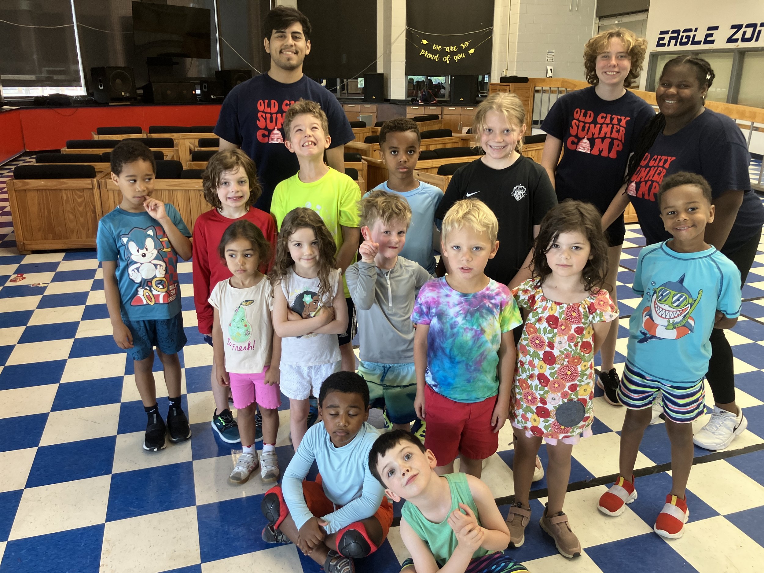 Group of children and two adults posing inside a gym or community center with blue and white checkered floor, wooden benches, and a banner that reads 'Old City Summer Camp' in the background.