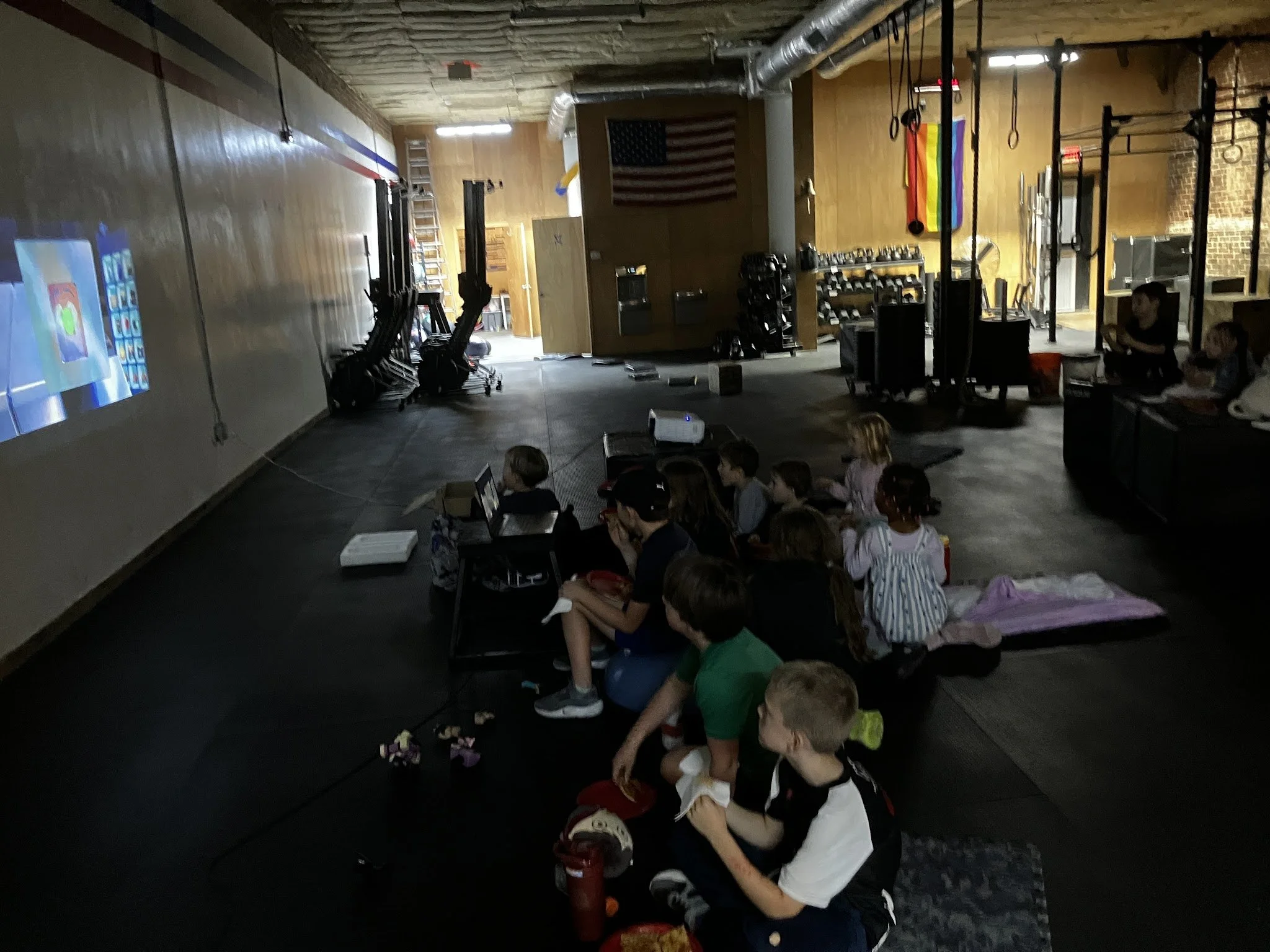 Children sitting on the floor in a gym or fitness space, watching a projected screen on the wall.