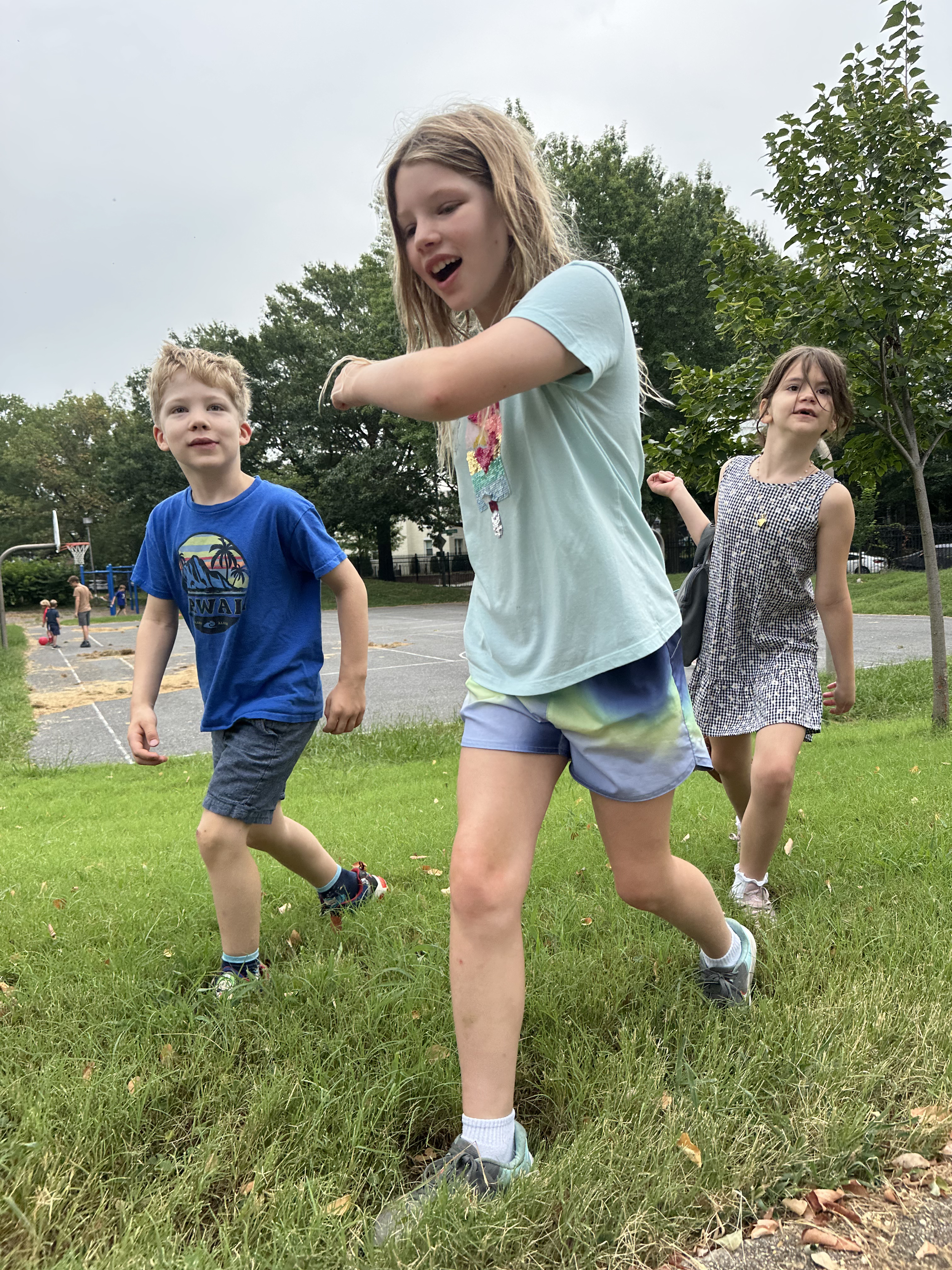Three children playing outside on a grassy area near a playground, with trees and overcast sky in the background.