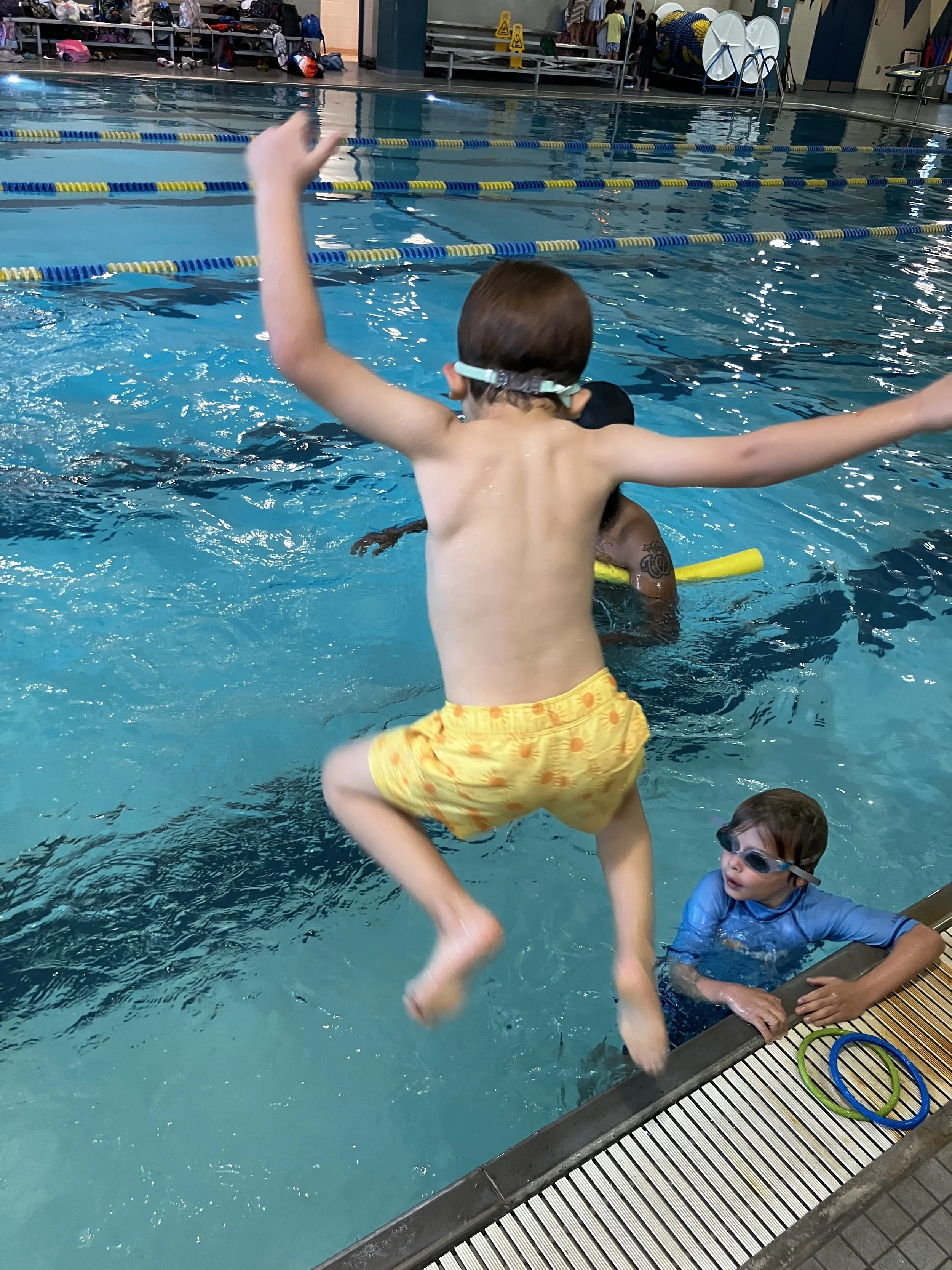 A young boy with brown hair jumping into a swimming pool, wearing yellow swim trunks with orange spots and goggles. Another child in blue swimwear and goggles is sitting at the pool's edge, watching.