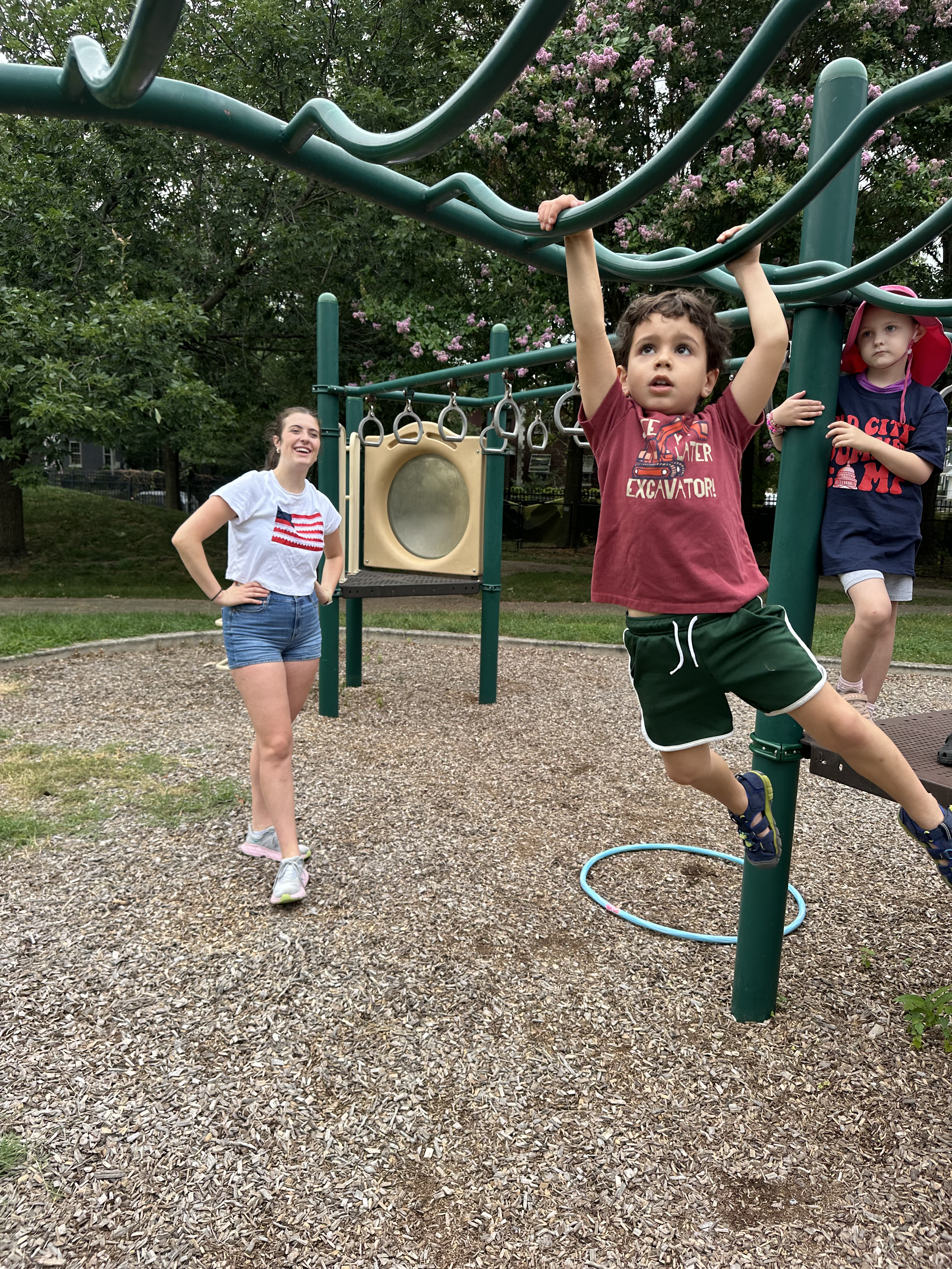 Children playing at a playground, with a boy hanging from a structure, a girl in the background, and a woman observing, all on a gravel surface with trees and flowering bushes in the background.