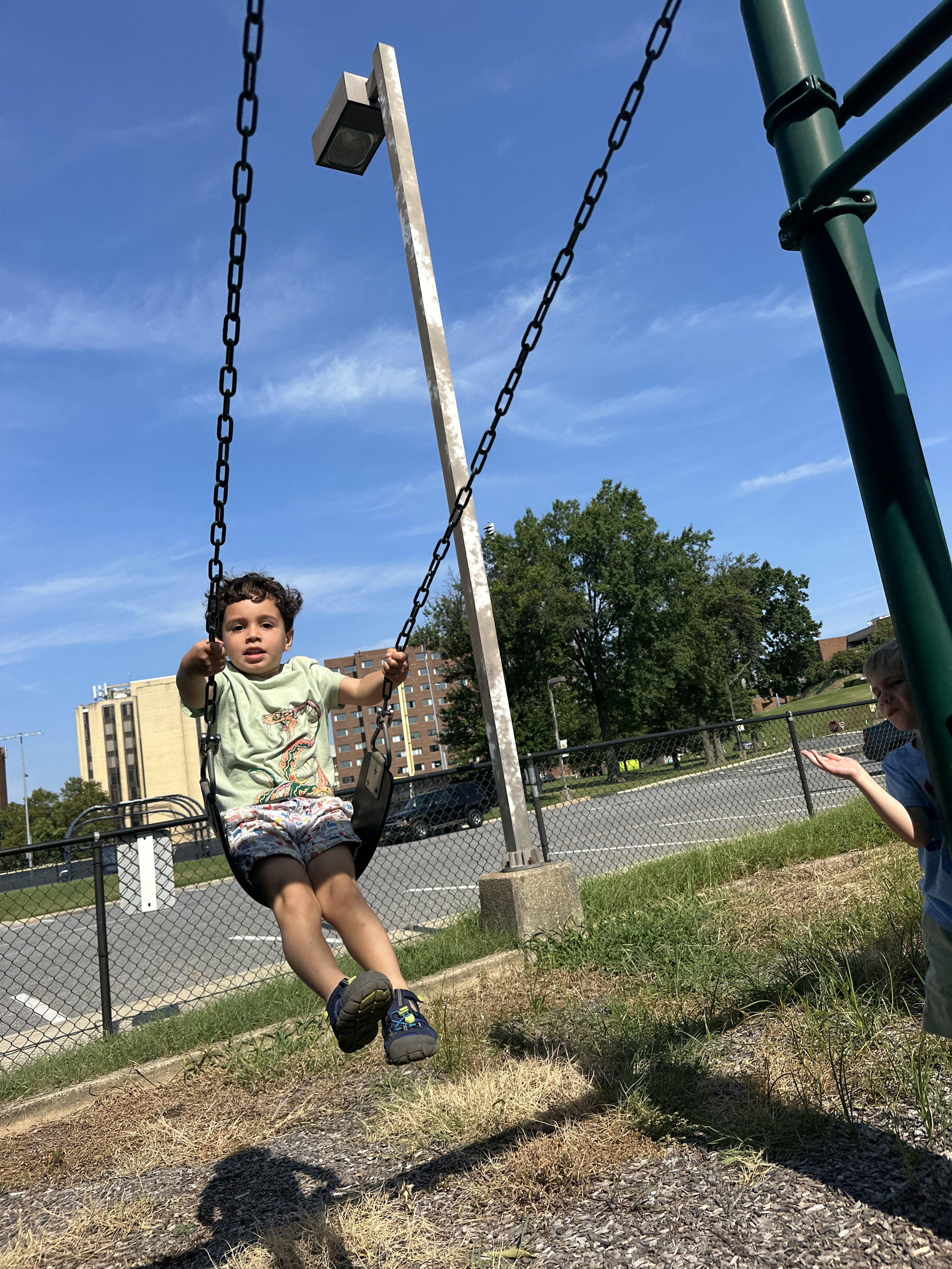 Child in a light green T-shirt with dinosaur design on swing at a playground on a sunny day.