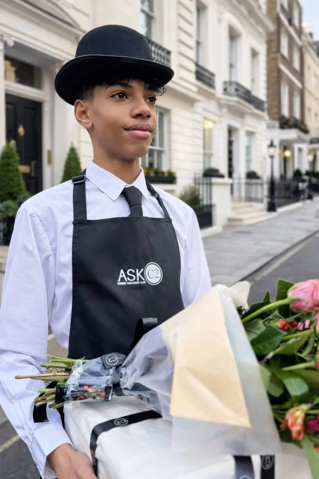 A young man in a white shirt, black apron, and bowler hat holding a bouquet of pink roses on a city street.