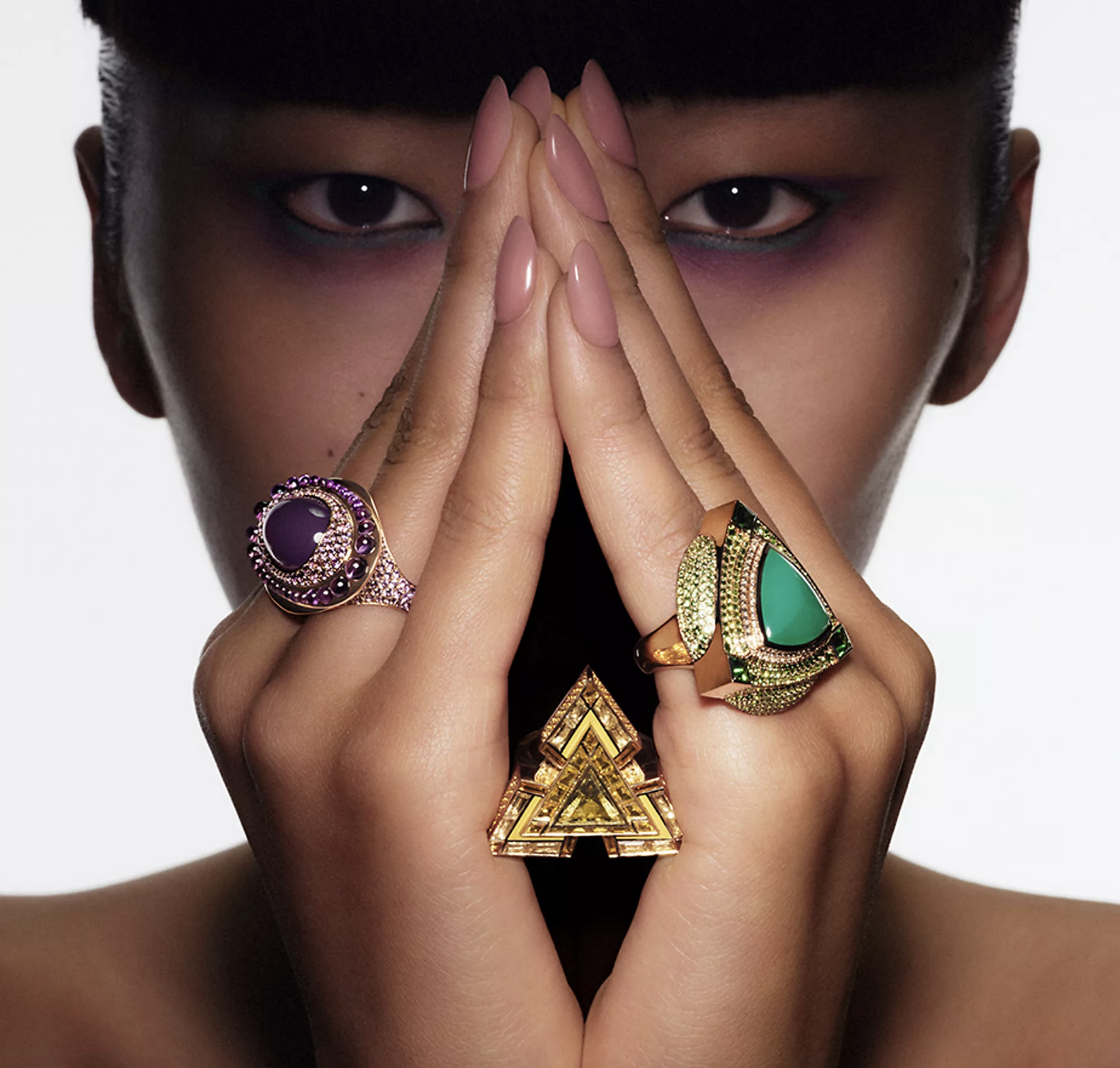 A close-up portrait of a person with hands pressed together in front of their face, showing multiple colorful rings on fingers, with a neutral background.
