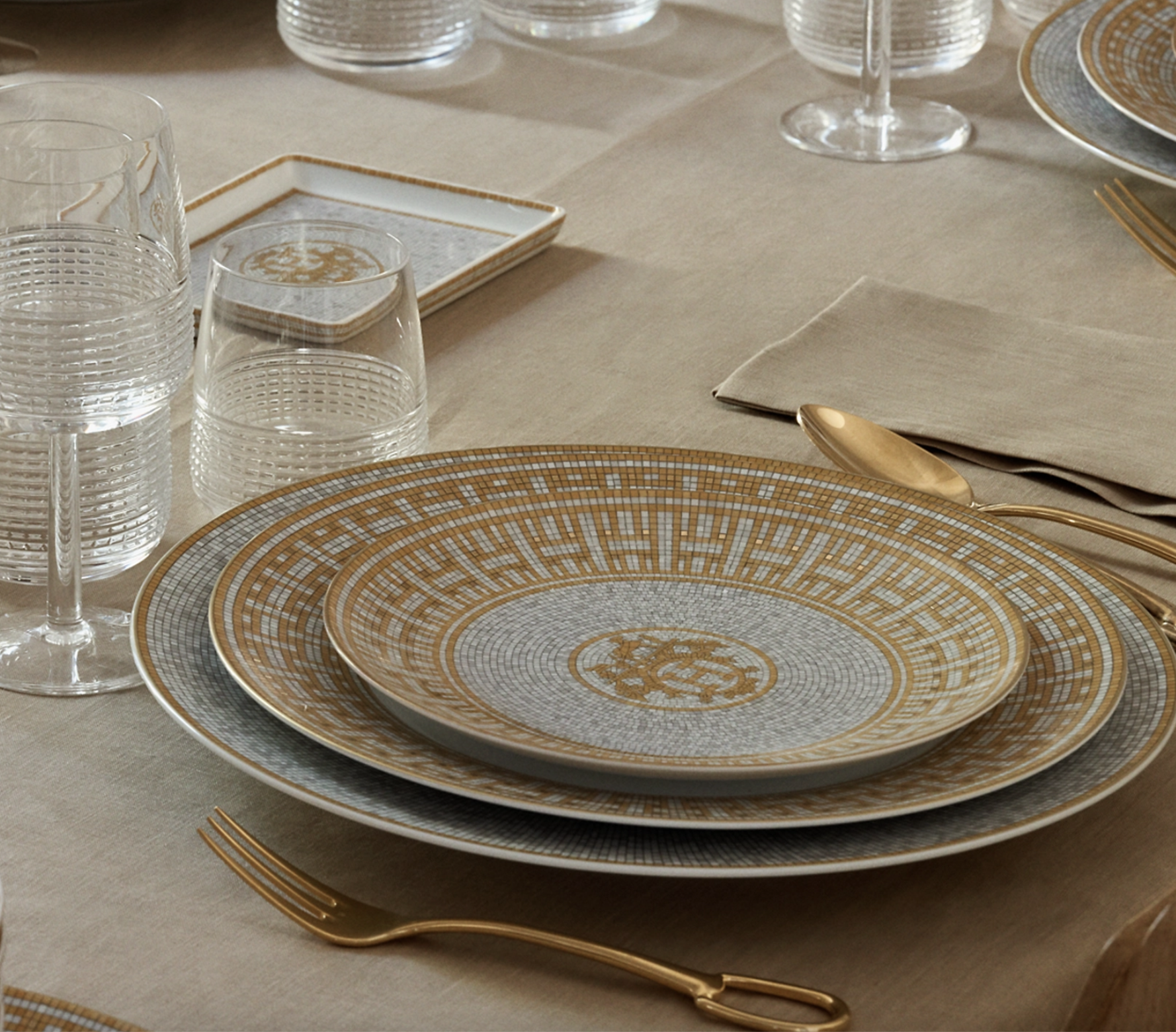 Elegant table setting with patterned plates, gold utensils, beige napkin, and clear glasses on a beige tablecloth.
