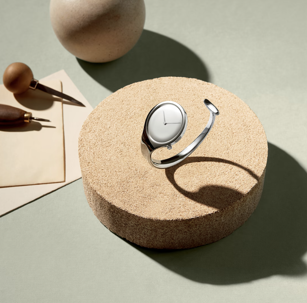 A modern watch with a sleek, rounded face and a metallic band rests on a circular textured platform. The scene includes a clay pot, a smaller round object, and two pens or styluses lying on a beige piece of paper, all set against a green surface.