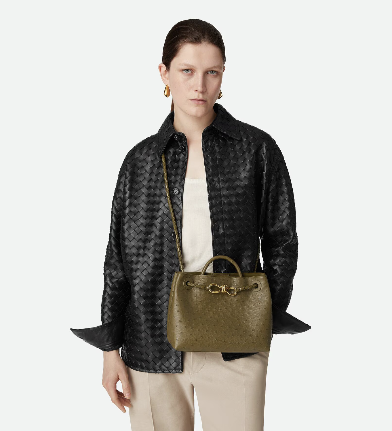 A woman with dark hair, wearing a black quilted leather jacket, a white top, and beige pants, holds an olive green handbag with a chain strap, standing against a plain background.