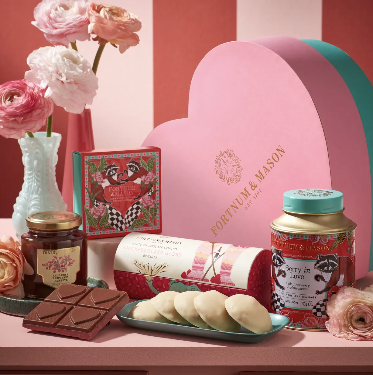 A collection of Valentine's Day gifts and treats, including a pink Fortune & Mason box, a jar of Rhubarb & Raspberry conserve, a chocolate bar, a box of biscuits, a container of Berry in Love tea, a plate of white chocolate-covered snacks, a vase wit