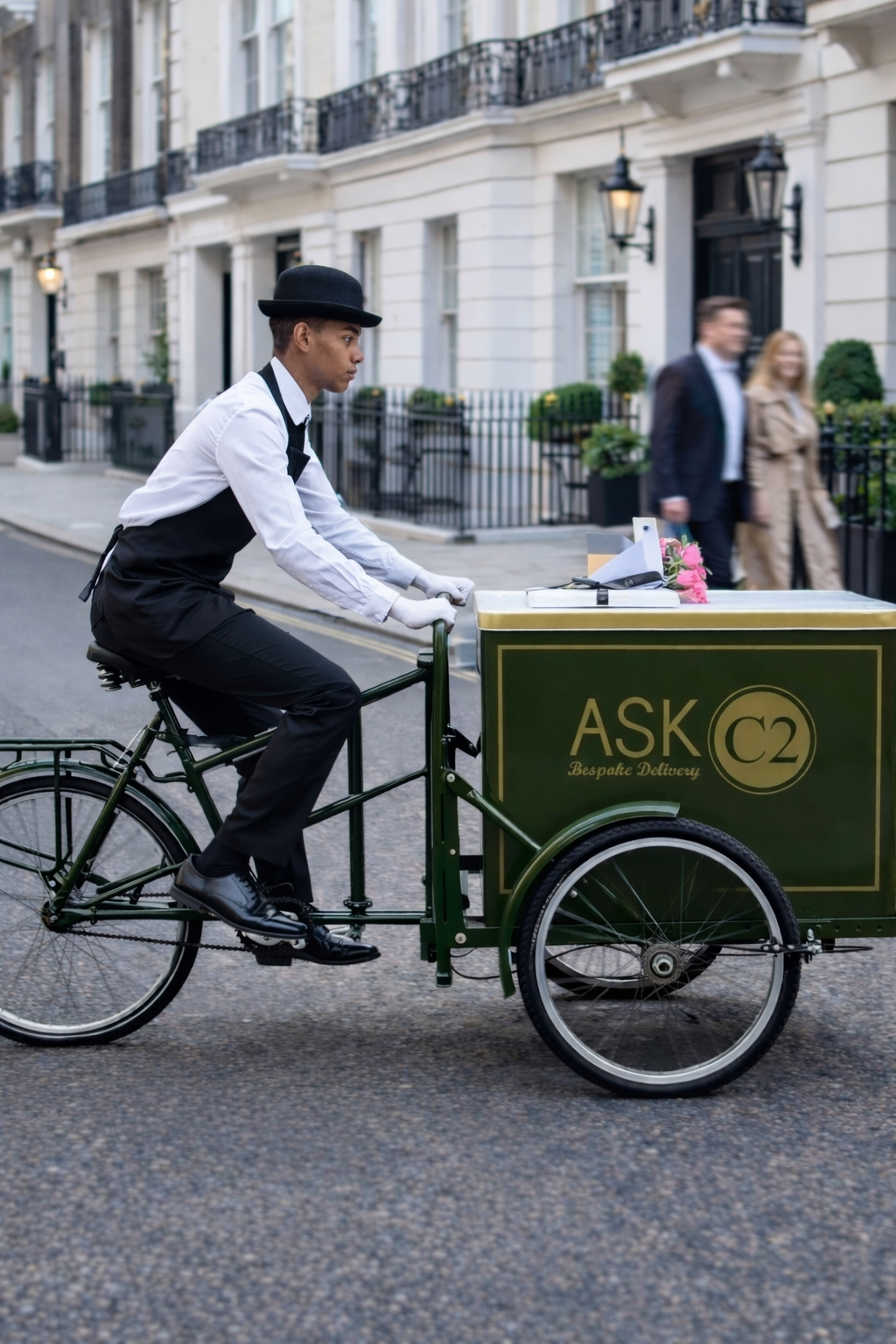 A young male delivery person rides a green  with a box labeled 'ASK C2 - Bespoke Delivery' on a city street in front of white apartment buildings with black railings. The rider is dressed in formal attire with a white shirt, black vest, and black bow