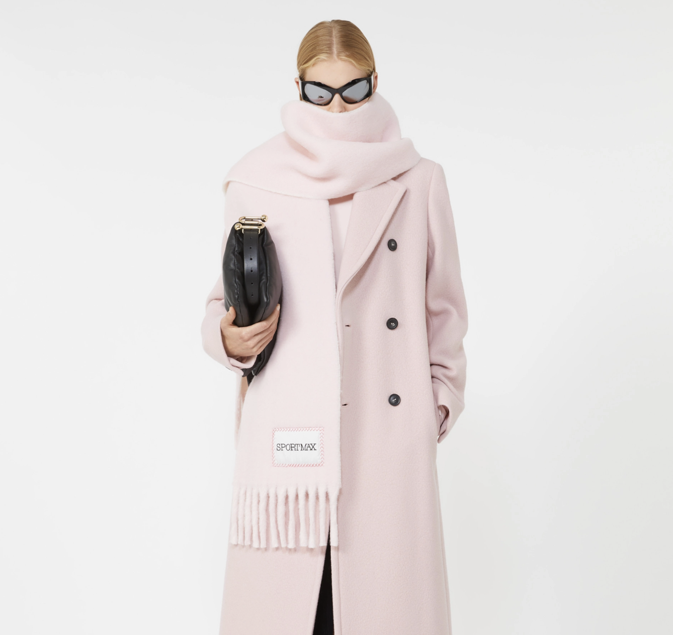 A woman wearing a pink coat, matching scarf, black sunglasses, and holding a black purse against a white background.