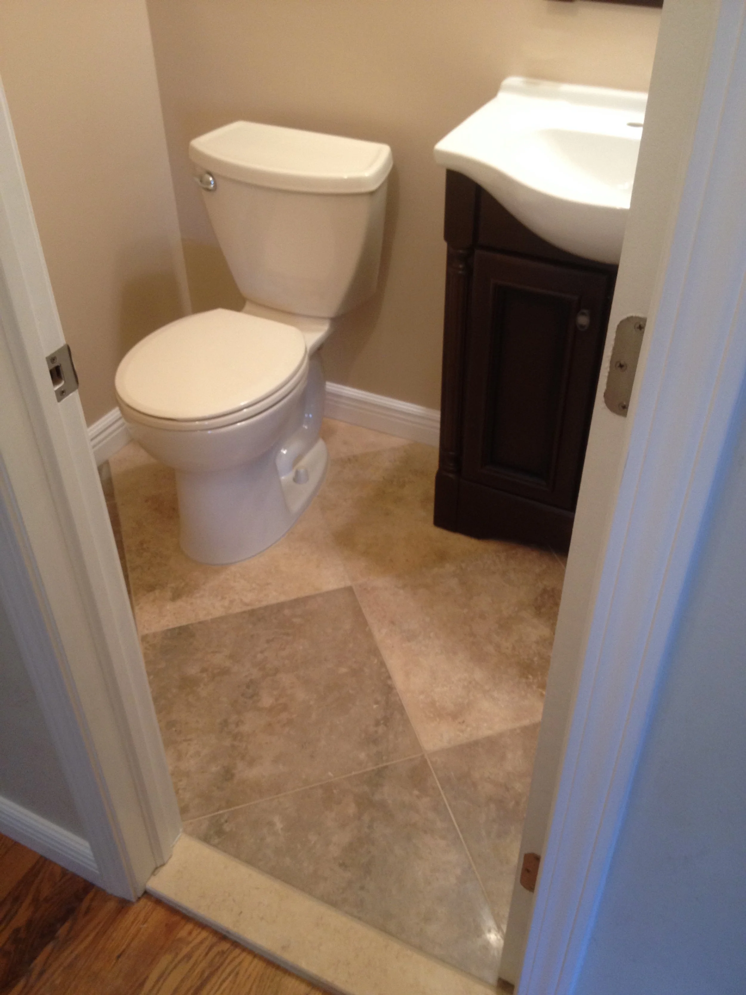 A small bathroom with a white toilet, a dark wooden cabinet with a white sink, beige walls, and tiled flooring.
