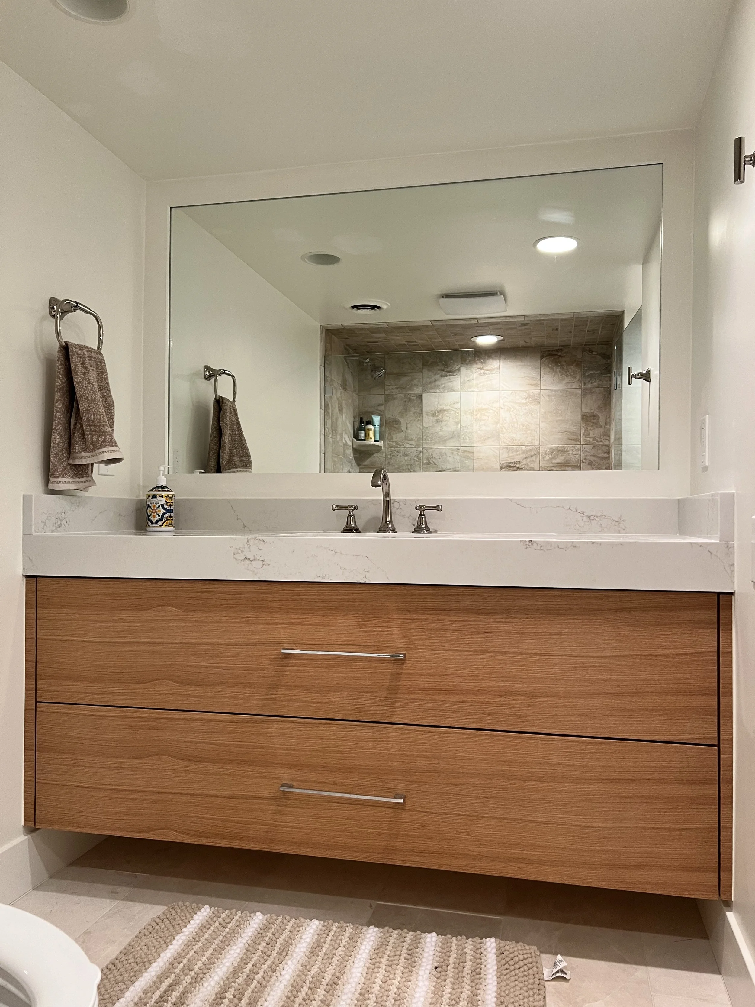 Bathroom vanity with wooden drawers, marble countertop with a sink, a large wall mirror, beige towels hanging on towel rings, and a soap dispenser, with a reflection showing a shower area with tiled walls and built-in shelves.