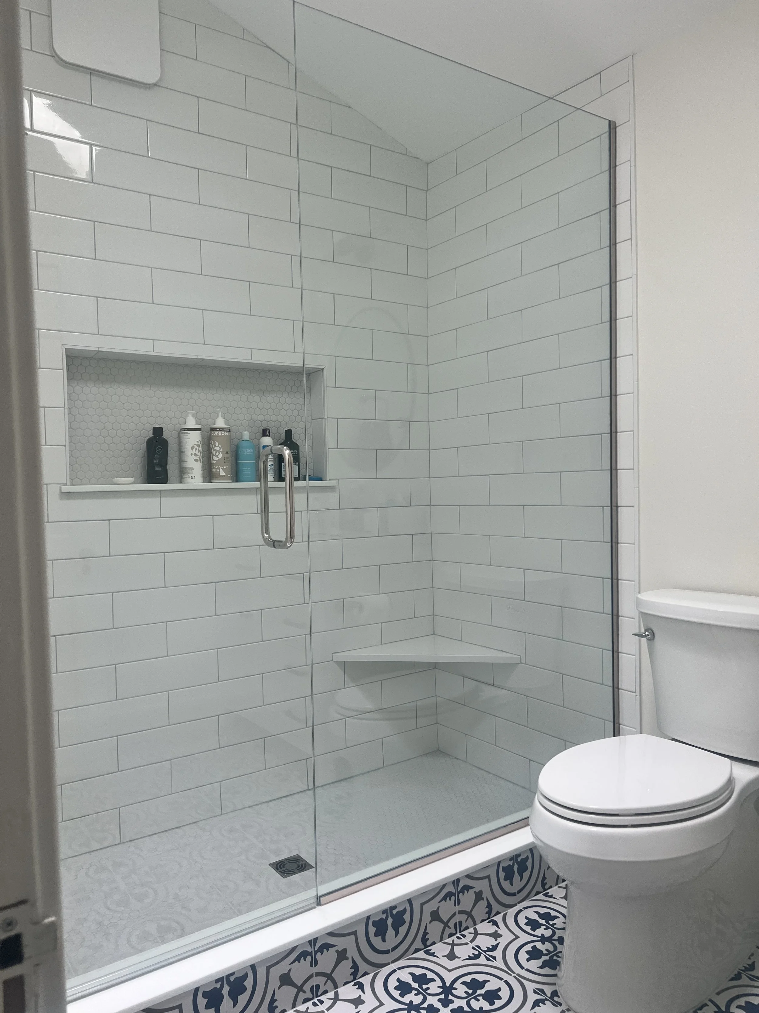 Modern bathroom with a glass shower enclosure, white tiled walls, built-in shower niche with toiletries, corner shower bench, and patterned floor tiles, next to a white toilet.