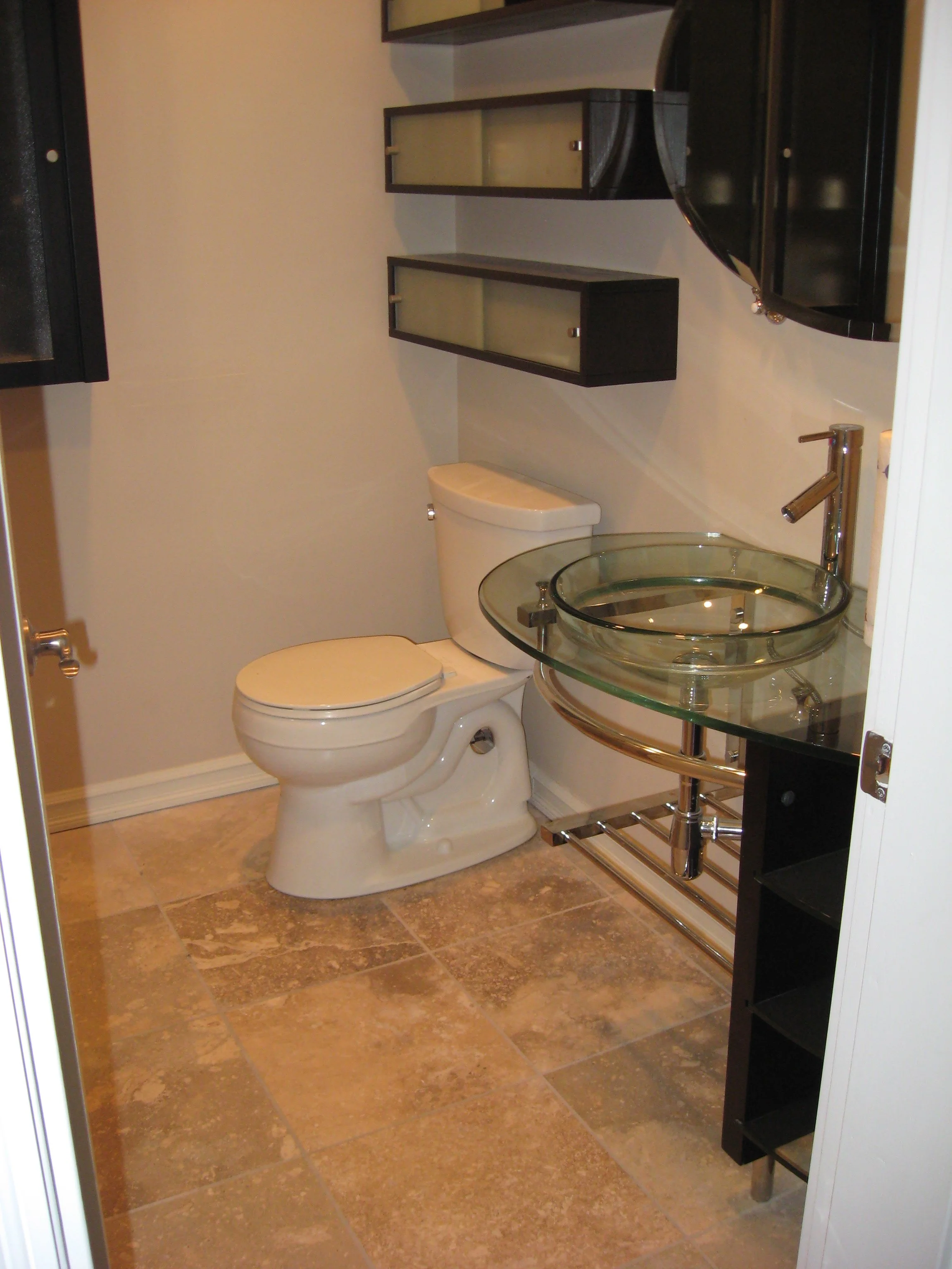Bathroom with a toilet, a glass vessel sink on a metal stand, and wall-mounted black shelves with glass doors.