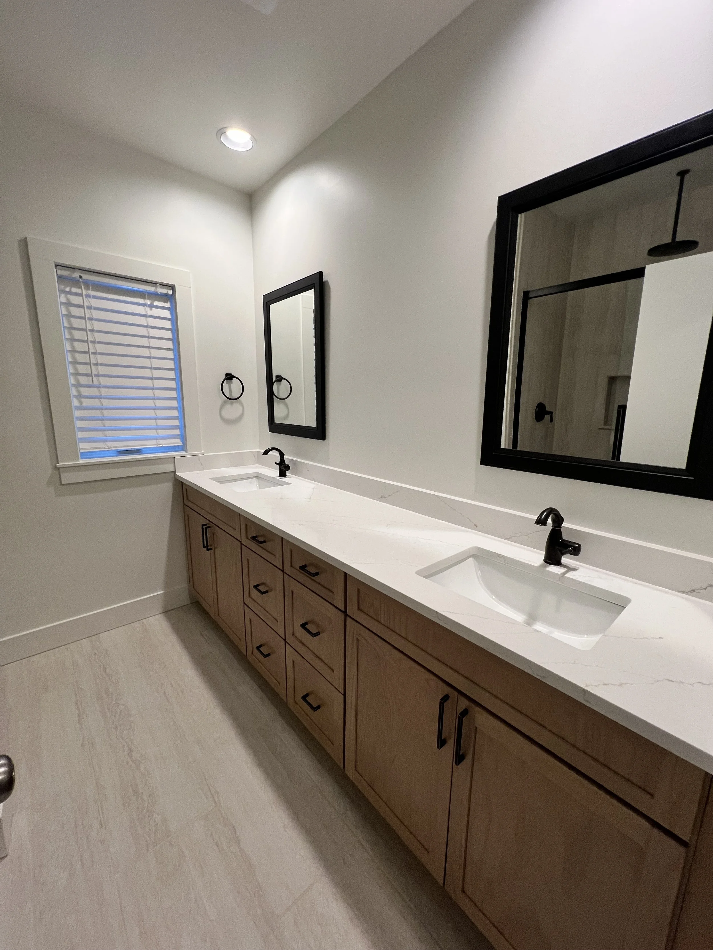 A modern bathroom vanity with dual sinks, white marble countertop, light wood cabinets, black fixtures, black-framed mirrors, a window with blinds, and a white wall with a recessed ceiling light.