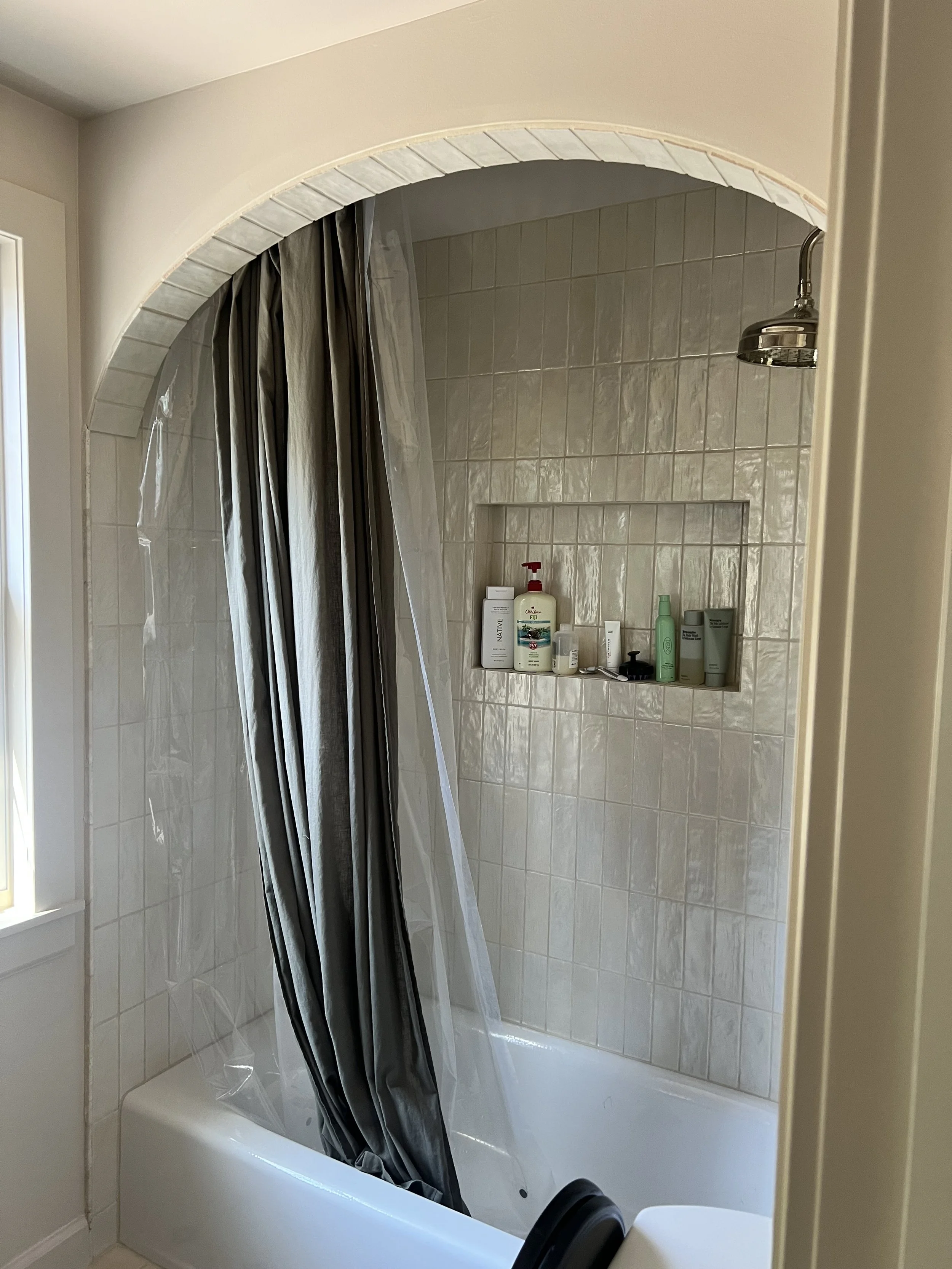Bathroom shower with beige tiled walls, a metal showerhead, and a built-in niche with personal care products. The shower has a clear plastic curtain and is next to a window.