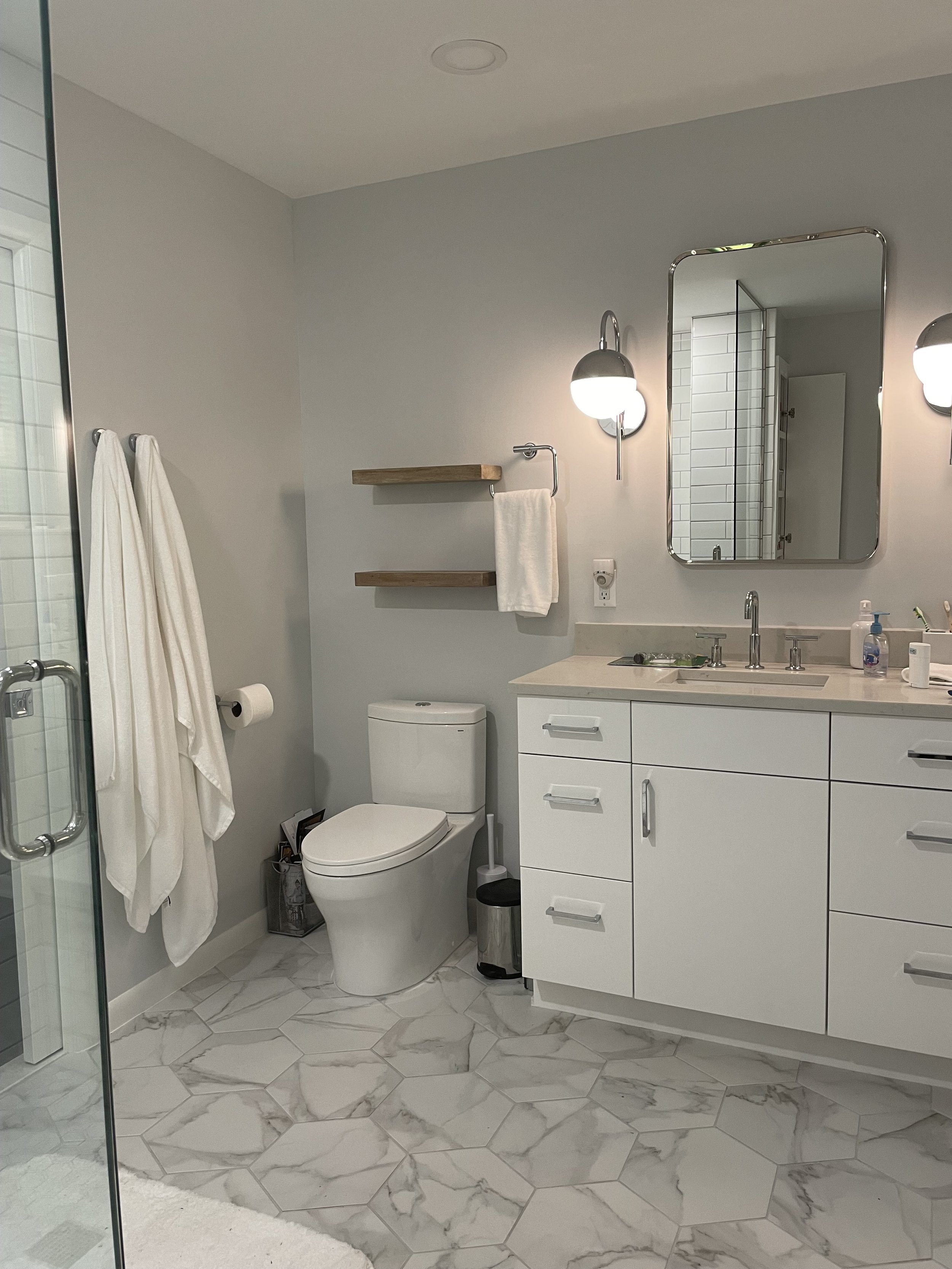 Modern bathroom with white vanity, marble hexagonal floor tiles, wall-mounted mirror, wall-mounted light fixtures, toilet, glass shower door, and wooden shelves with towels.