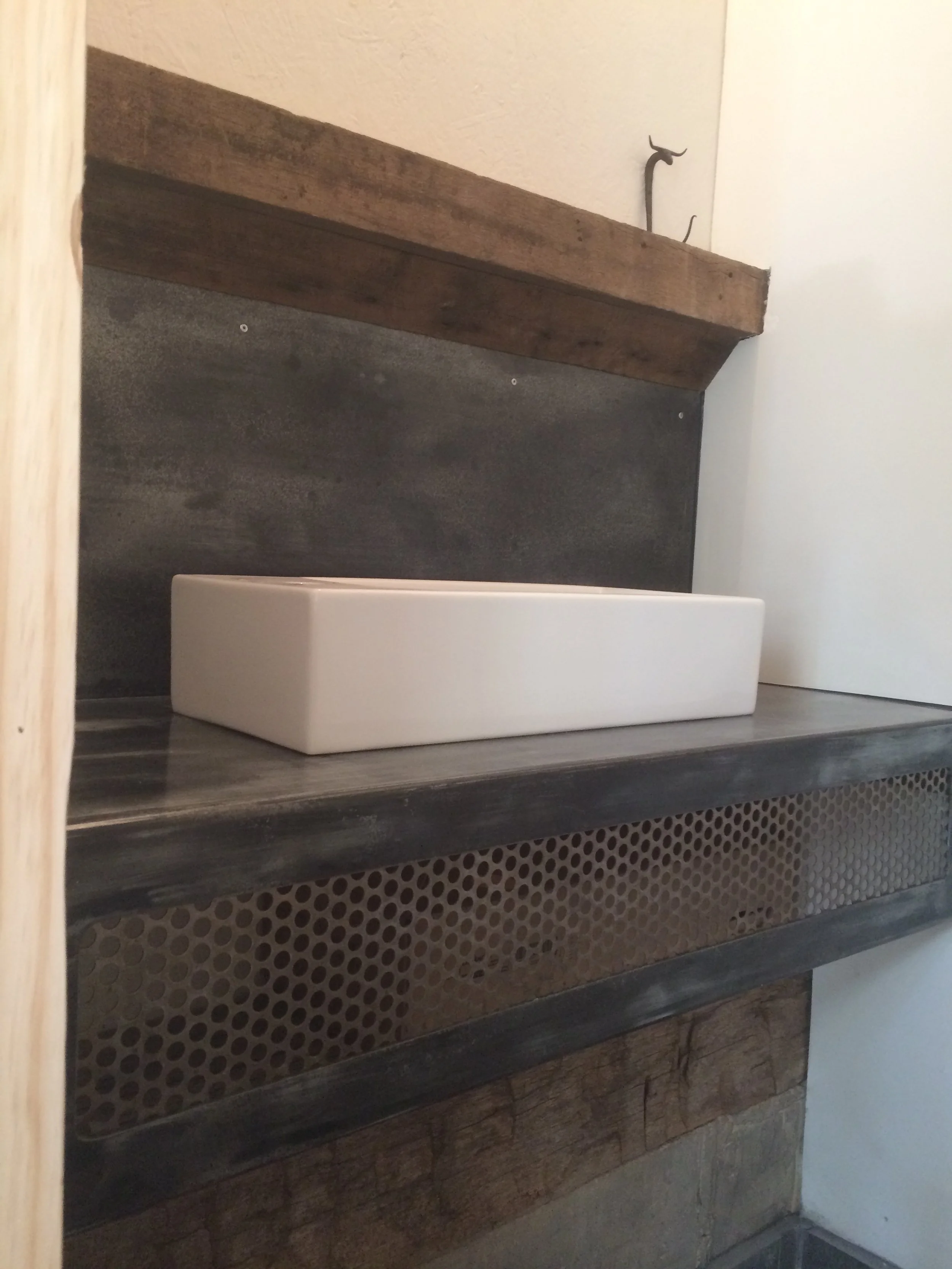 Close-up of a modern bathroom sink with a rectangular white basin on a rustic wooden vanity with a metal perforated drawer front, against a dark textured backsplash and wooden shelf above.