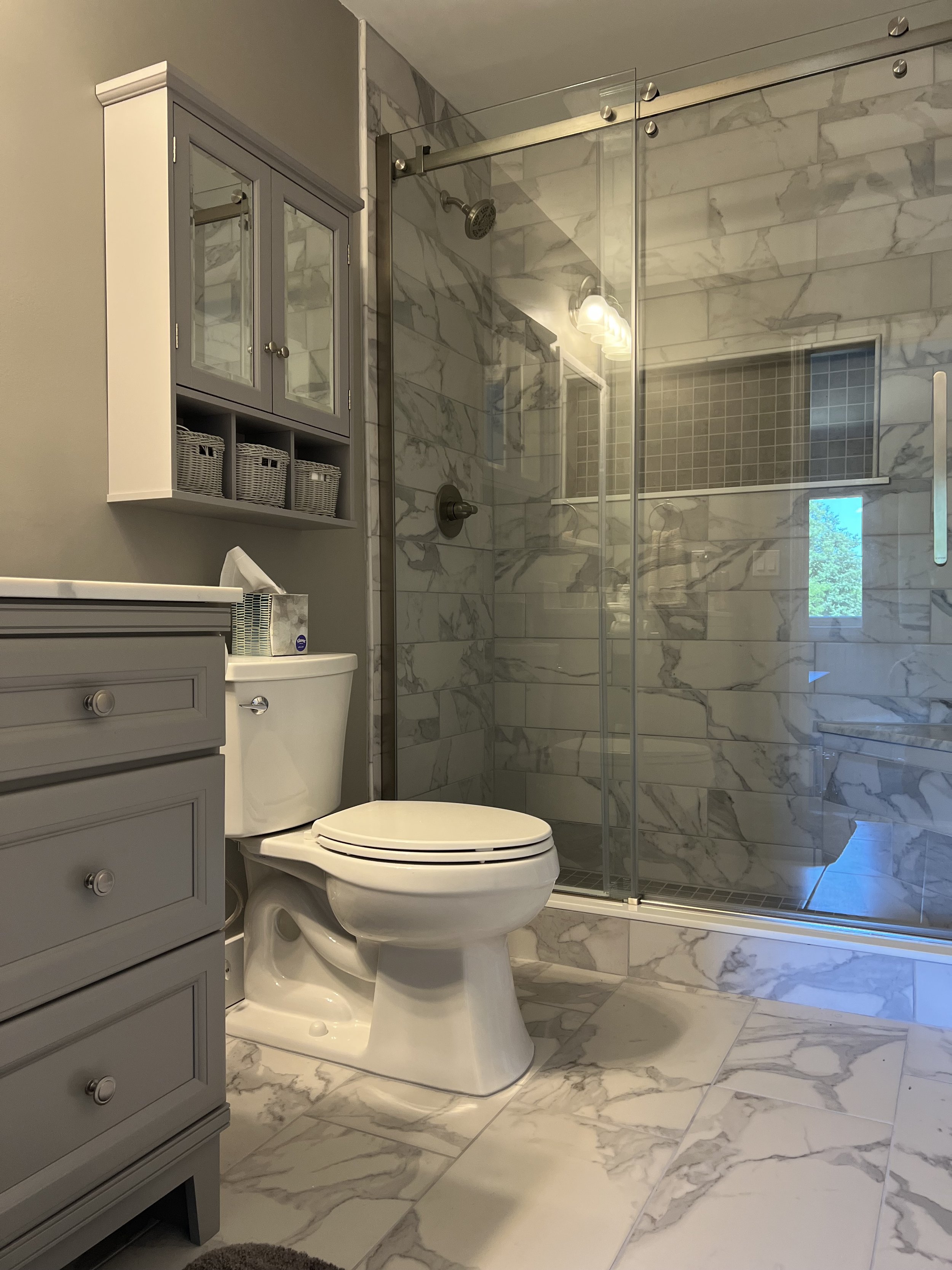 A bathroom with gray and white marble tile on the floor and shower walls, a white toilet, a gray cabinet with drawers, a wall-mounted gray cabinet with glass doors and baskets inside, and a glass-enclosed shower with a window showing outdoor greenery