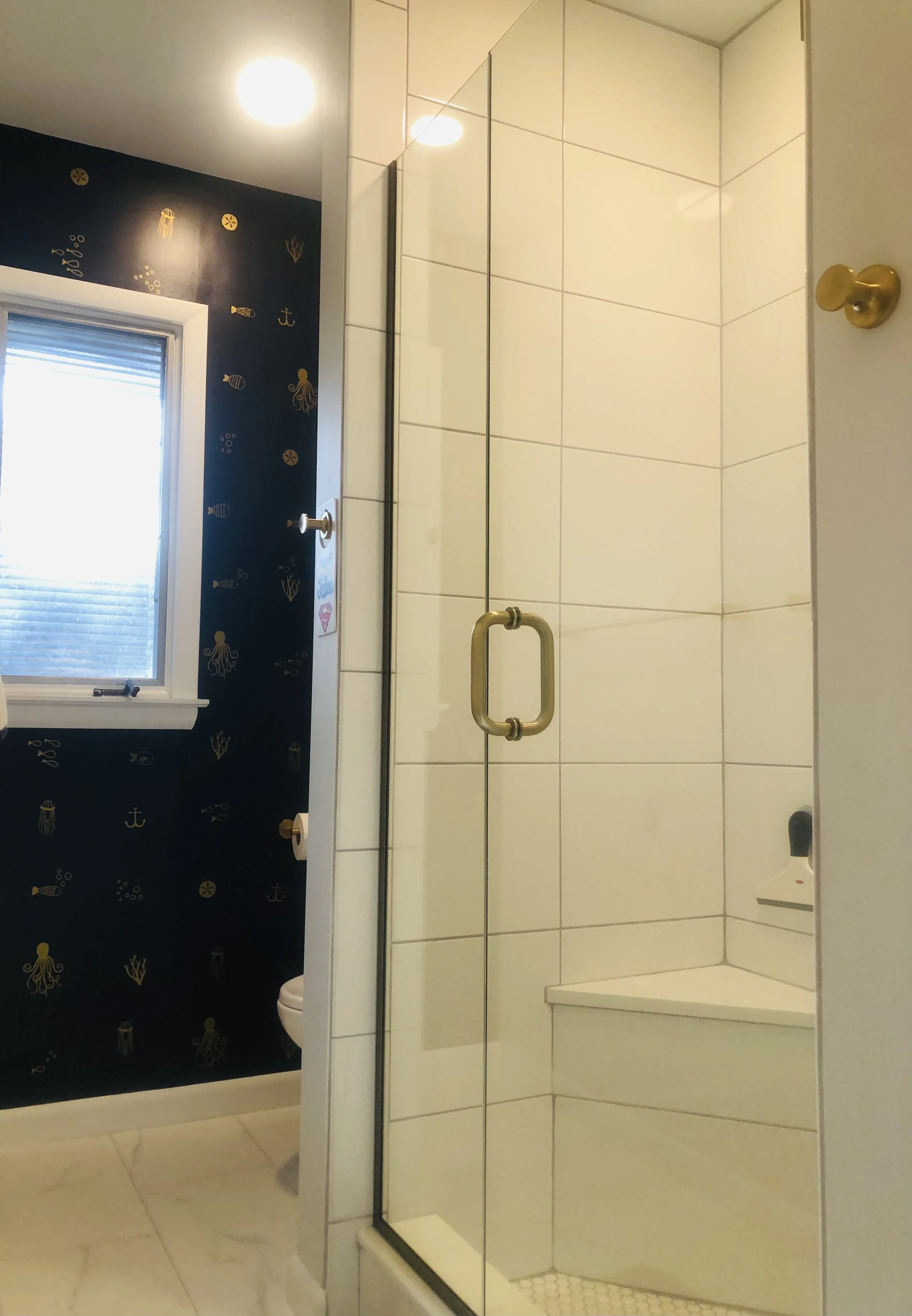 A bathroom with a glass shower enclosure, white tiled walls, a brass handle, a brass hook on the door, a small corner seat inside the shower, and a navy blue wall with nautical-themed decals. There is a window with blinds and a light fixture on the c