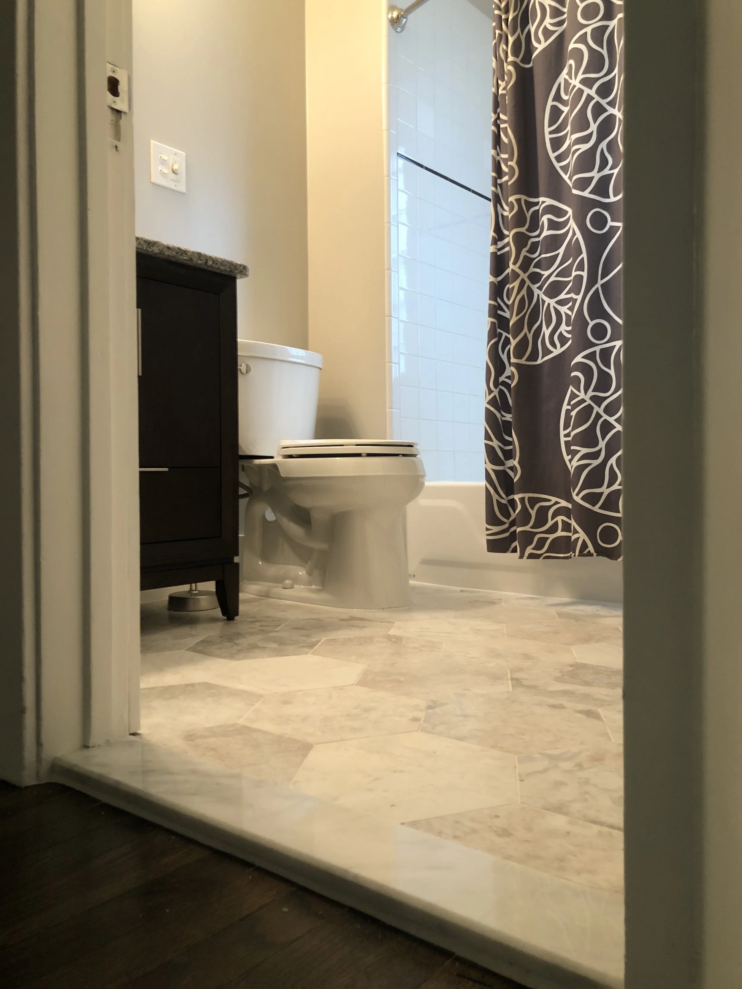 View of a bathroom with a toilet, a dark wooden cabinet, a shower with a patterned shower curtain, and beige tiled flooring.