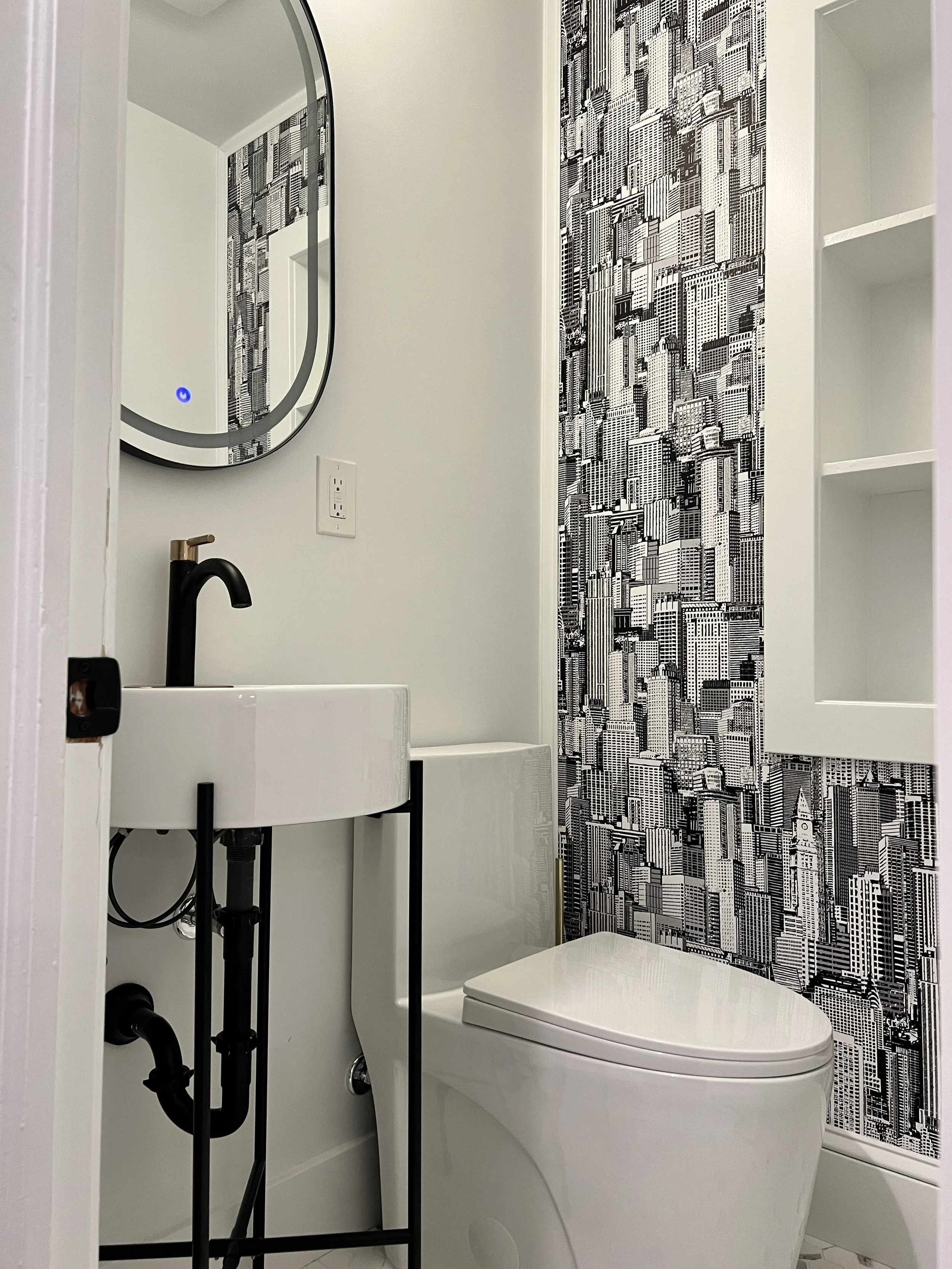 Small modern bathroom with black faucet, oval mirror, black cityscape wallpaper behind toilet, and built-in white shelves.
