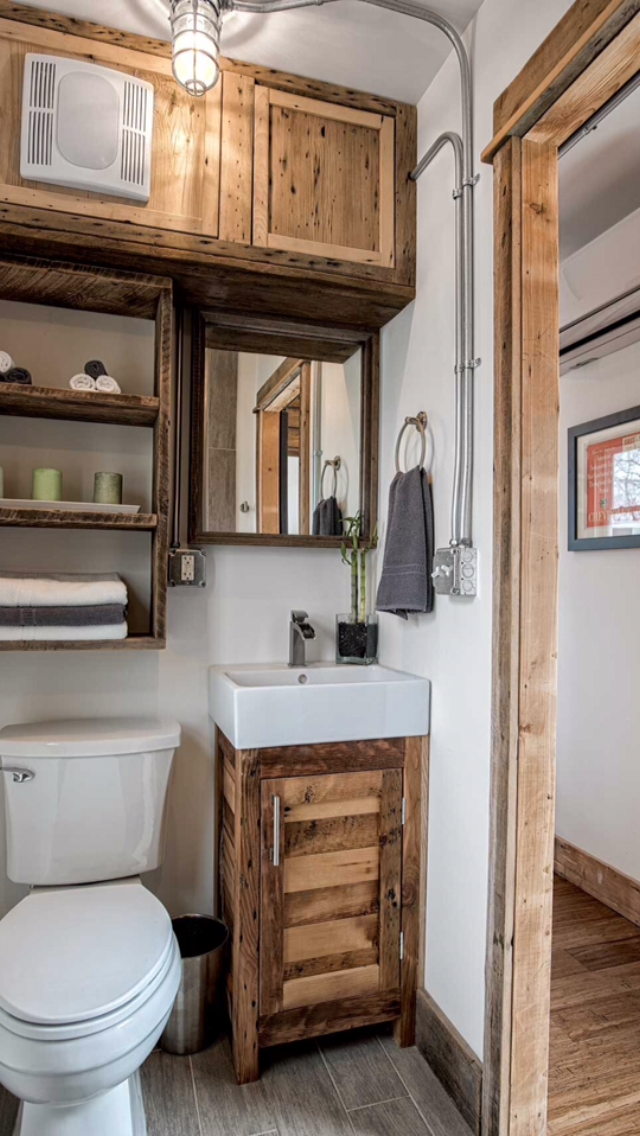 Small bathroom with wooden accents, a white toilet, a wooden cabinet with a sink, a mirror, an open shelf with towels and candles, and a vent on the wall.