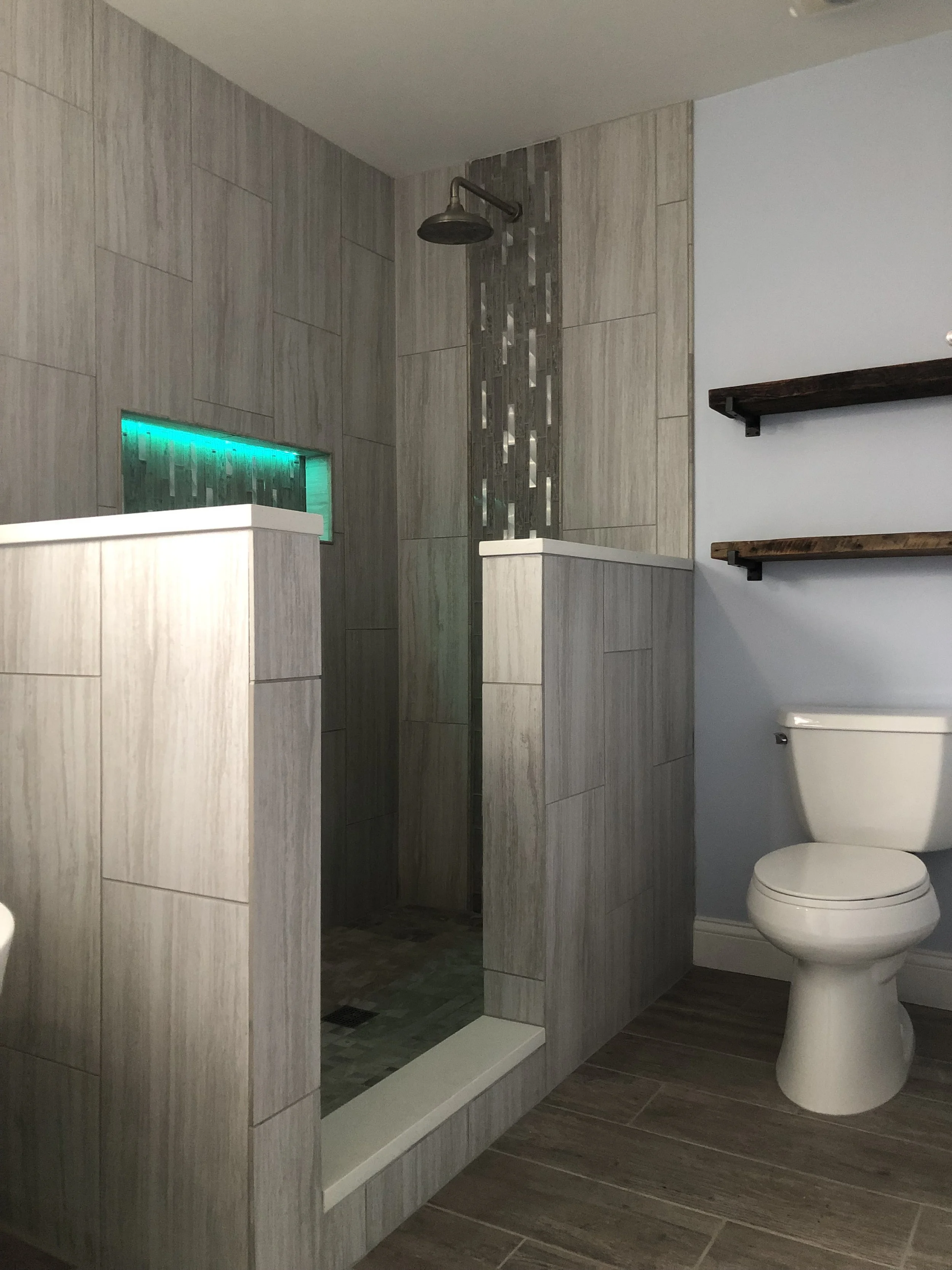 A modern bathroom with a walk-in shower featuring gray tiles, a rain shower head, and a glass block accent with colored lighting. There is a toilet and wooden shelving on the wall. The floor is wood-look tile.