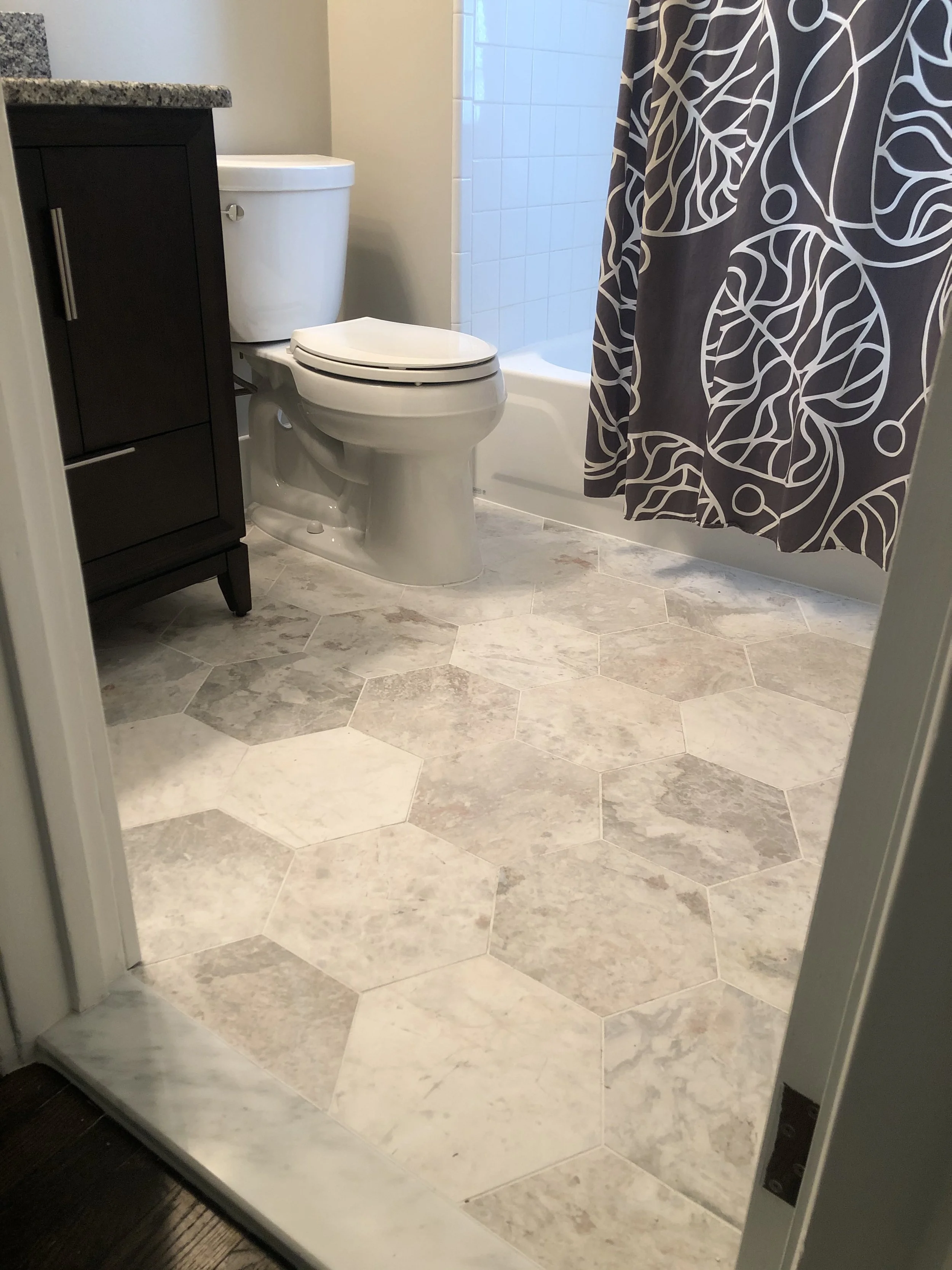 A small bathroom with a white toilet, a dark wood vanity, a shower with a patterned curtain, and hexagonal marble floor tiles.