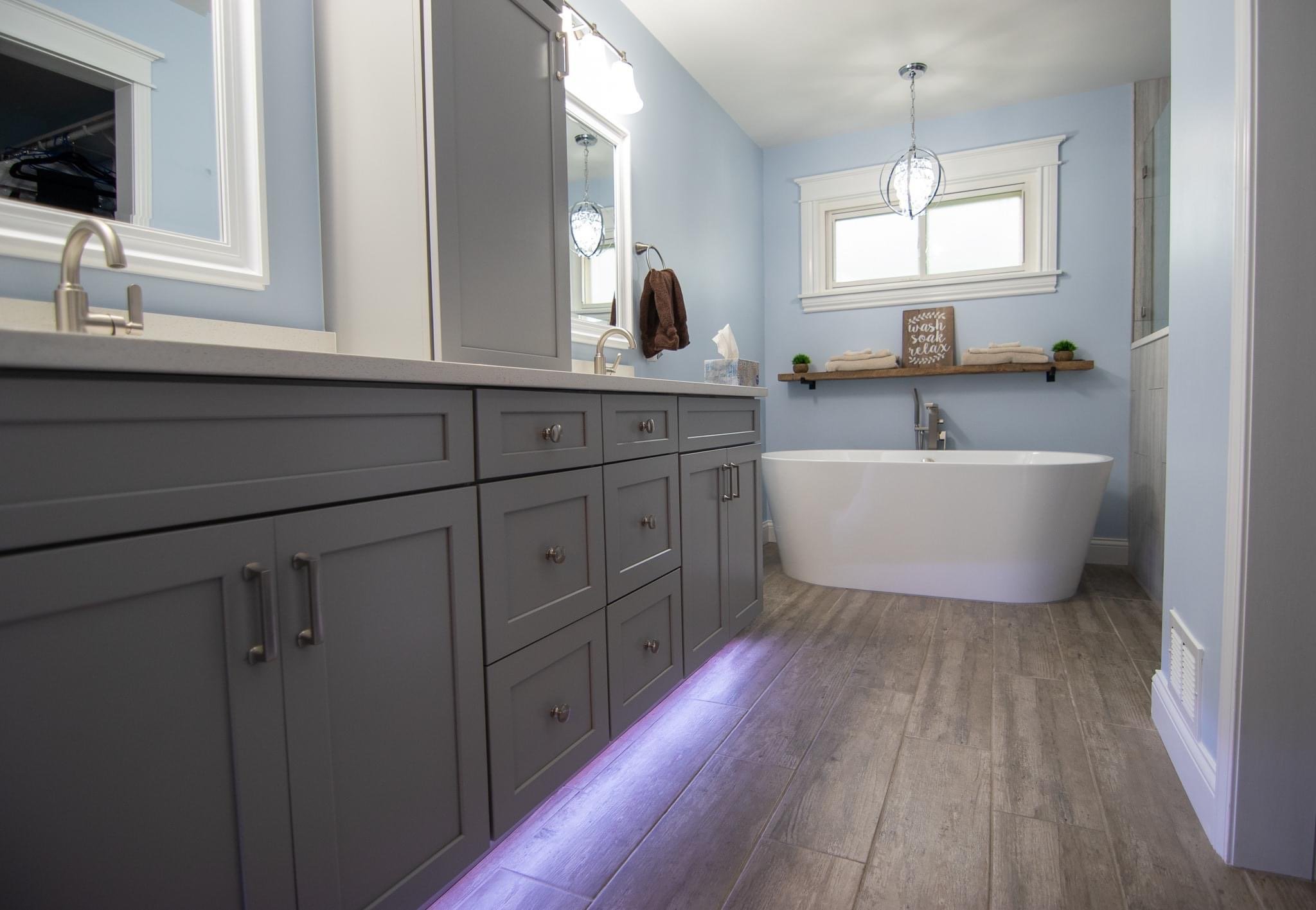 Modern bathroom with blue walls, gray vanity, white bathtub, wooden shelf, and pendant light fixtures.