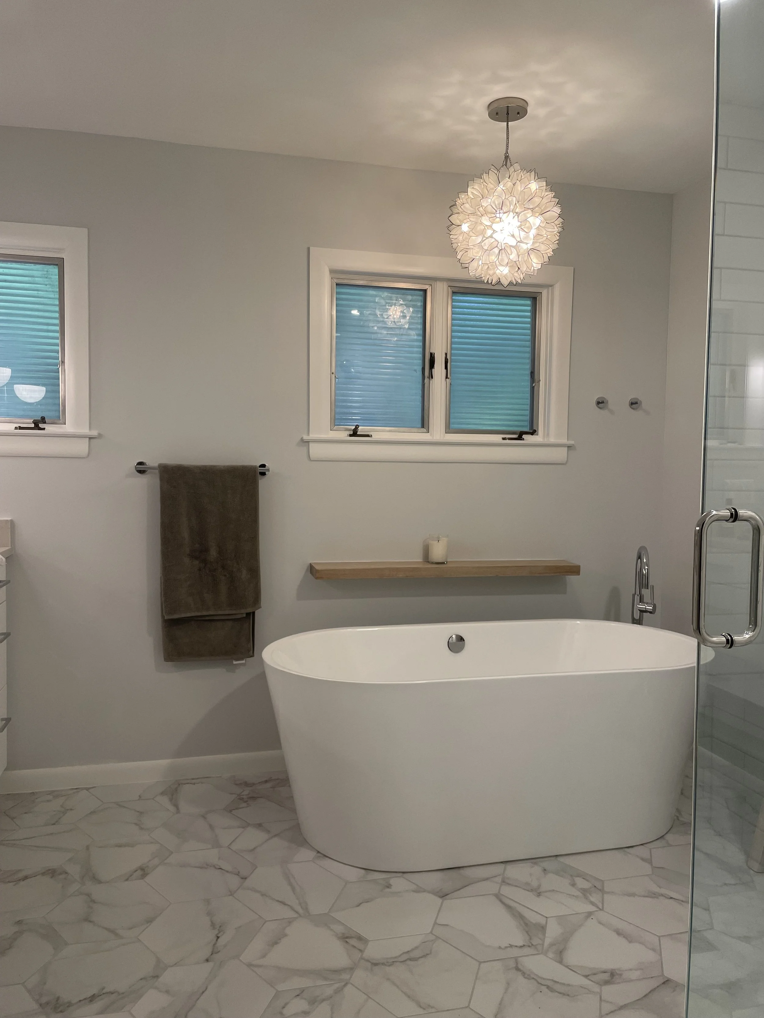 A modern bathroom with a white bathtub, marble tile floor, two small windows with closed blinds, a wooden shelf with a candle, a textured pendant light, and a towel hanging on a bar.