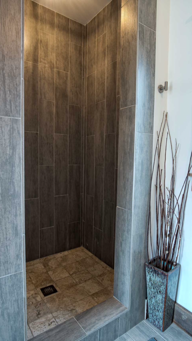 A small shower with beige and brown tiled walls and floor, and a drain at the center of the shower floor. There is a decorative vase with sticks beside the shower.