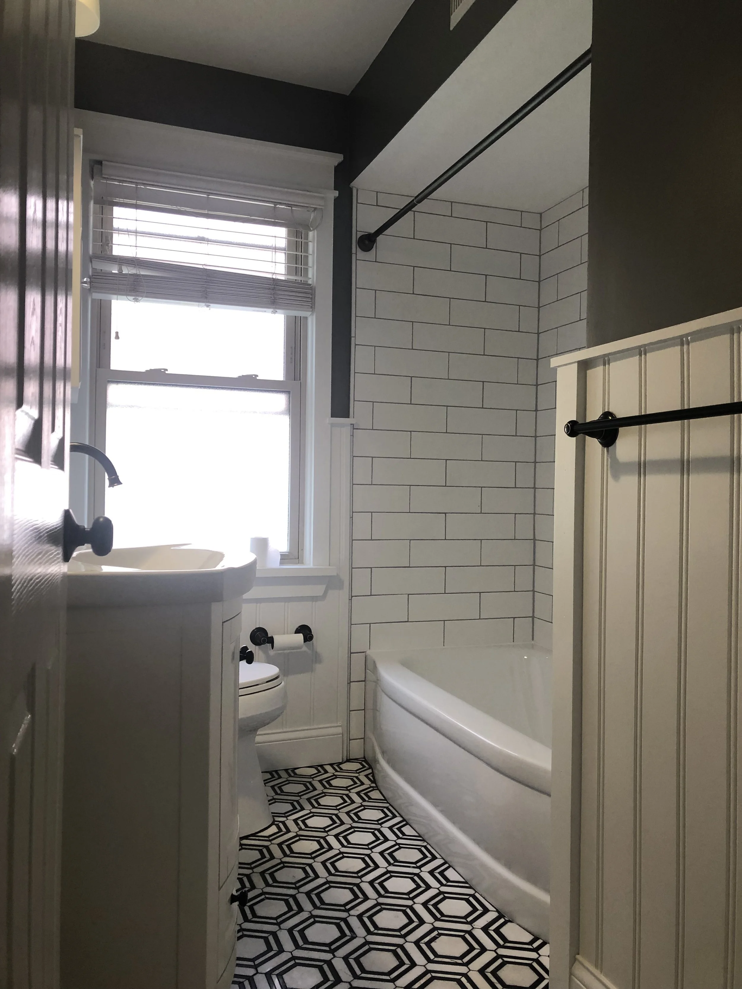 Small bathroom with black and white patterned floor, white bathtub, porcelain toilet, small white sink with black fixtures, window with blinds, dark walls, and white beadboard paneling.