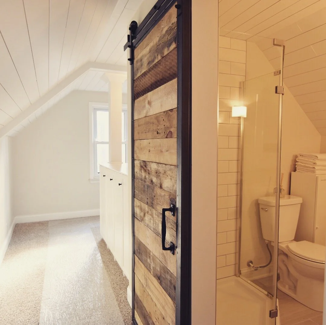 A rustic sliding barn door made of reclaimed wood planks in front of a small bathroom with a toilet and shower in an attic space with sloped ceiling and small window.
