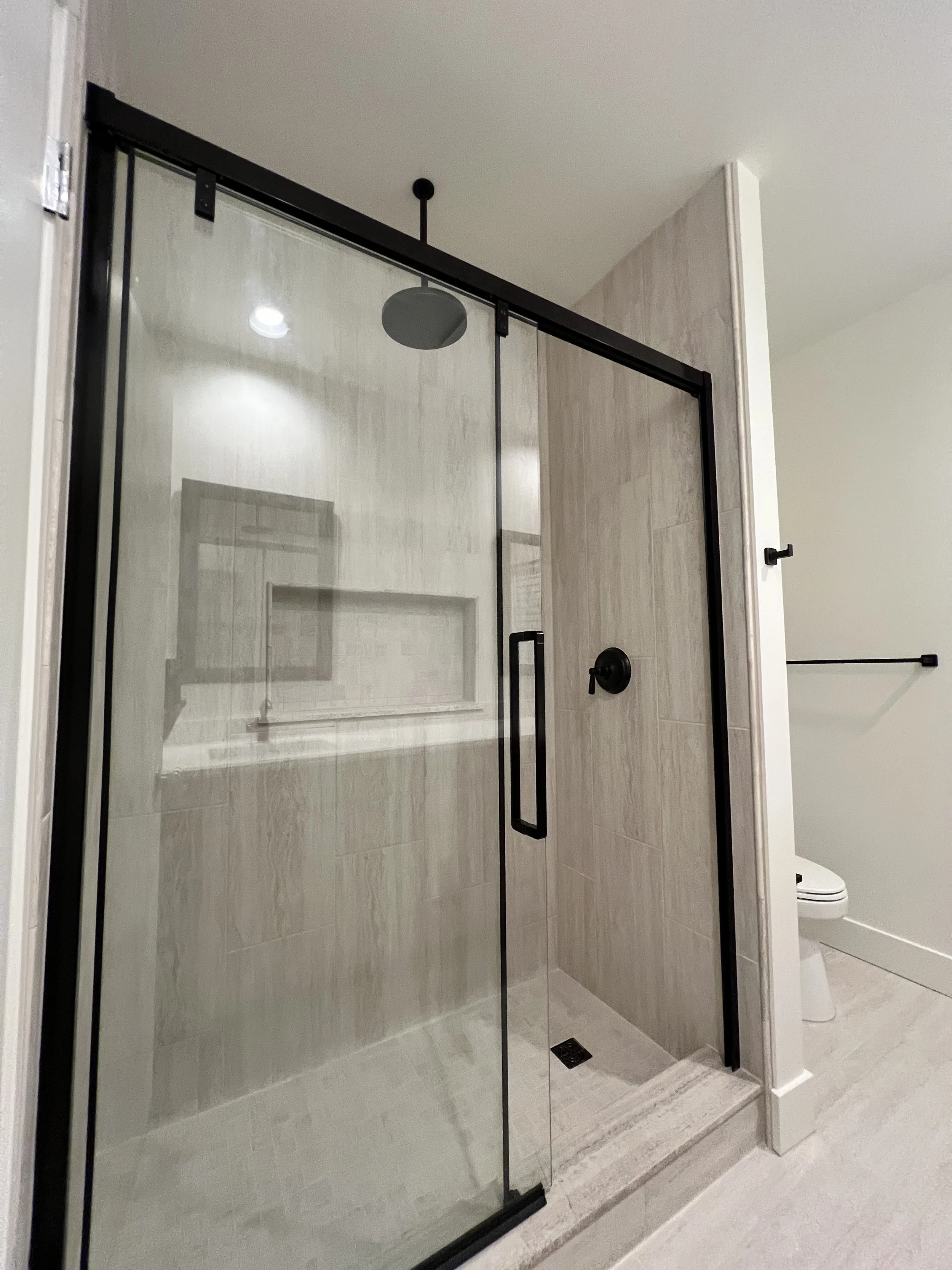 Bathroom shower with glass door, black hardware, beige tiled walls and floor, and a white toilet visible in the background.