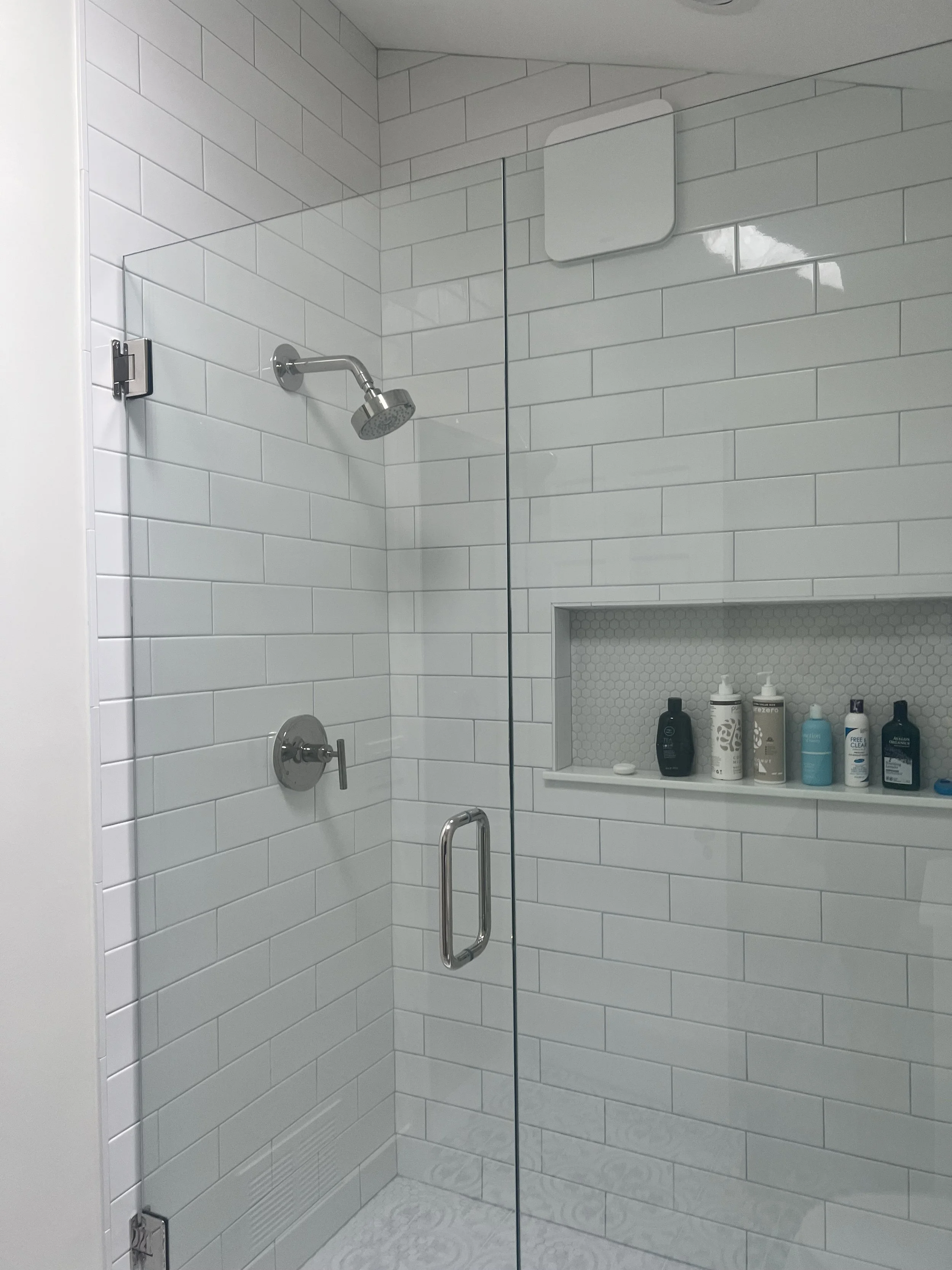 A glass shower door with metal handle in a bathroom shower area. The shower has white tiles and a built-in shelf with various bottles of toiletries.