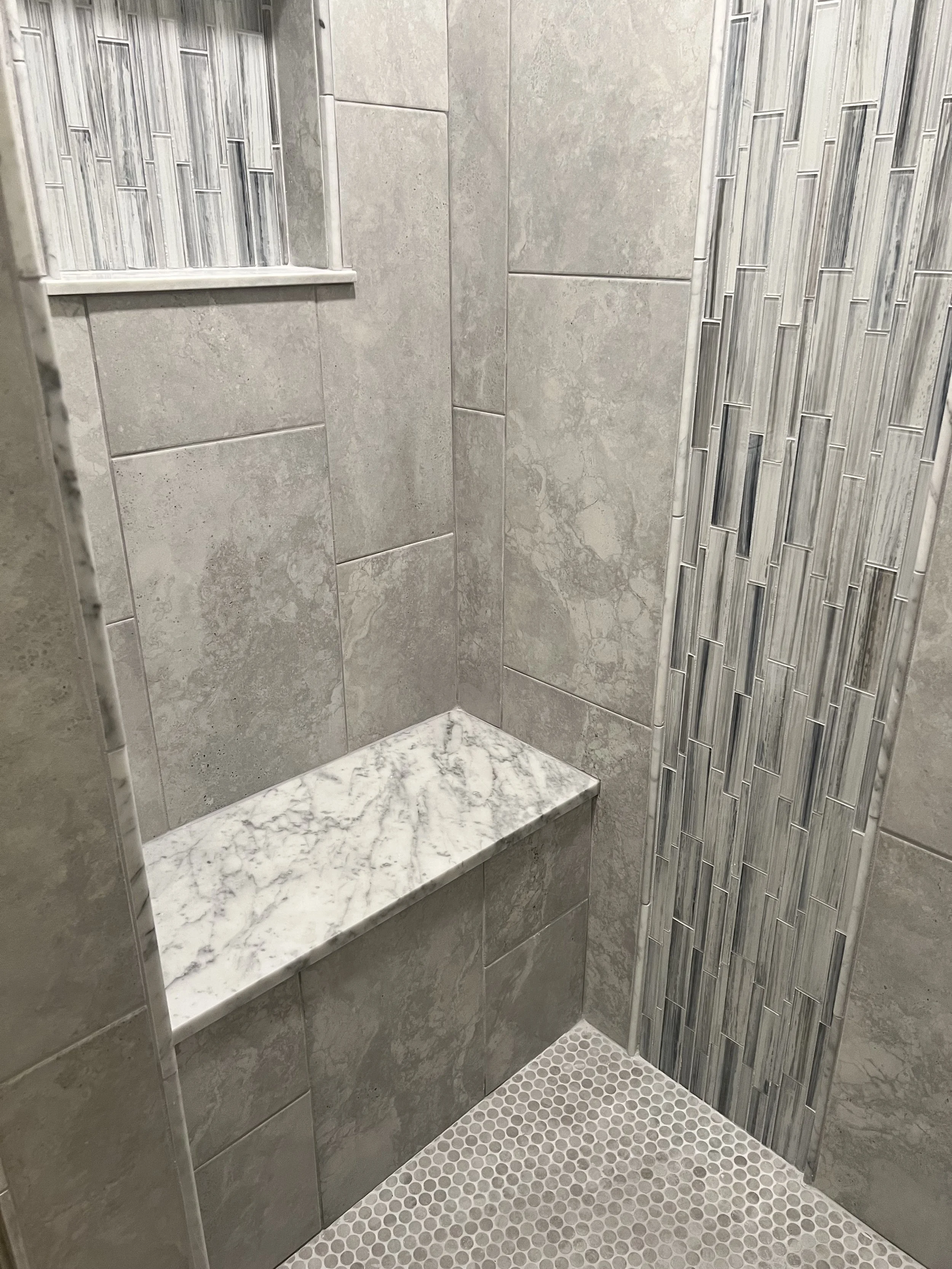 Close-up view of a modern shower corner with grey ceramic tiles, a built-in marble bench, and decorative tile accent wall in a shower stall.