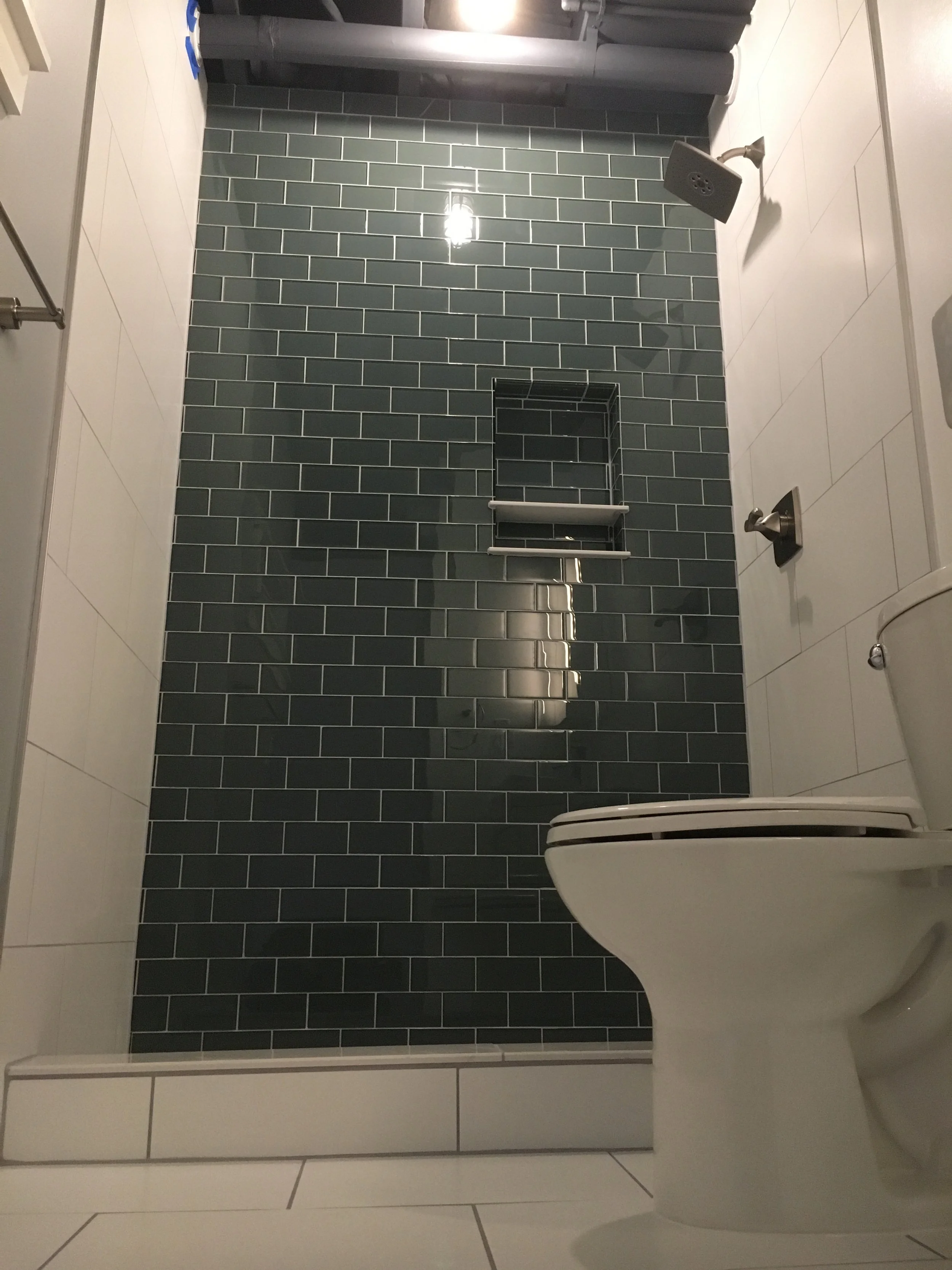 Modern bathroom shower area with dark gray tiles on the shower wall, a showerhead, a small built-in shelf with a towel, and a toilet in the lower right corner.