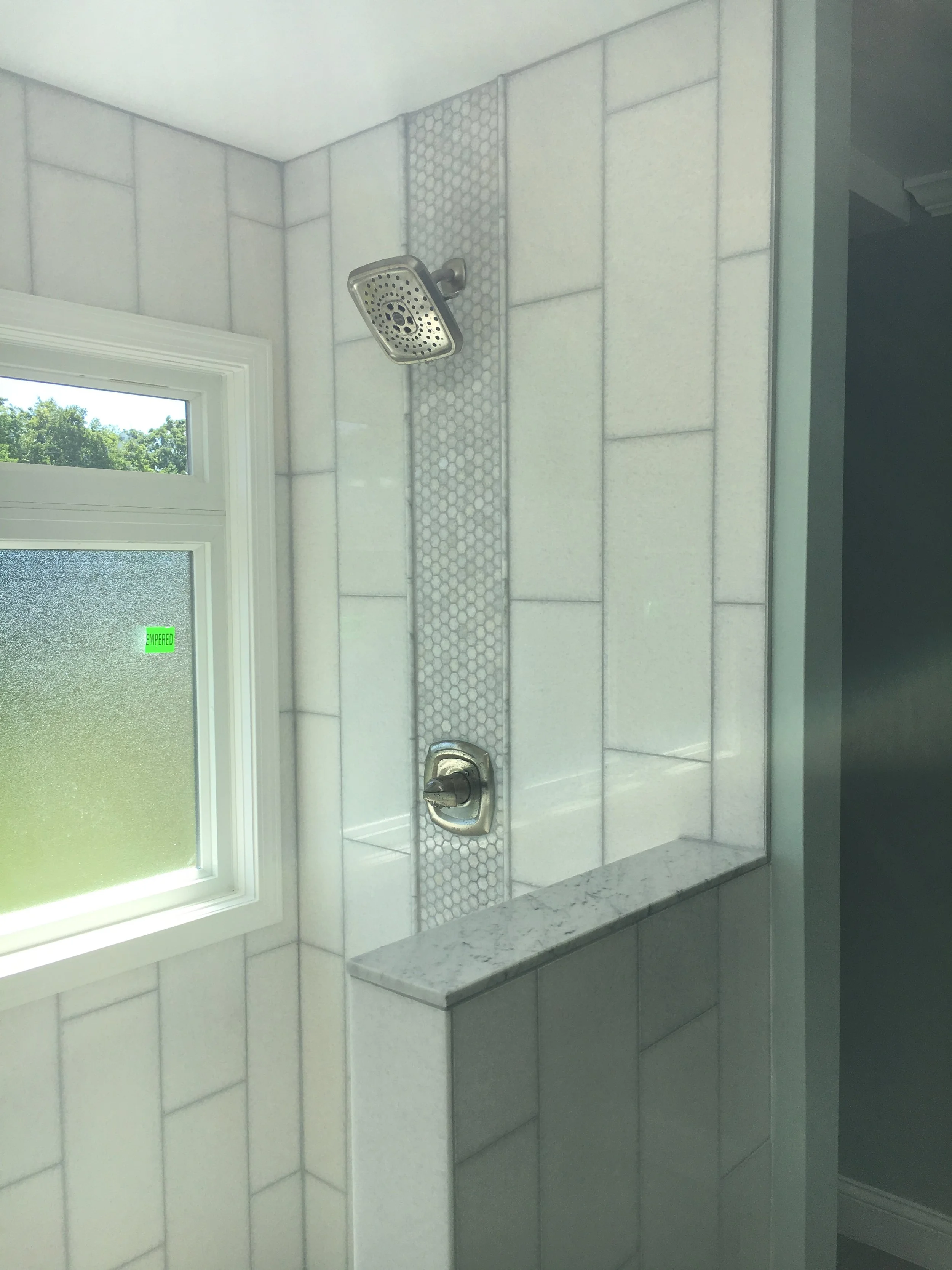 Shower area with a window, chrome showerhead, and a chrome control handle on a tiled wall, with a narrow strip of small hexagonal tiles running vertically.