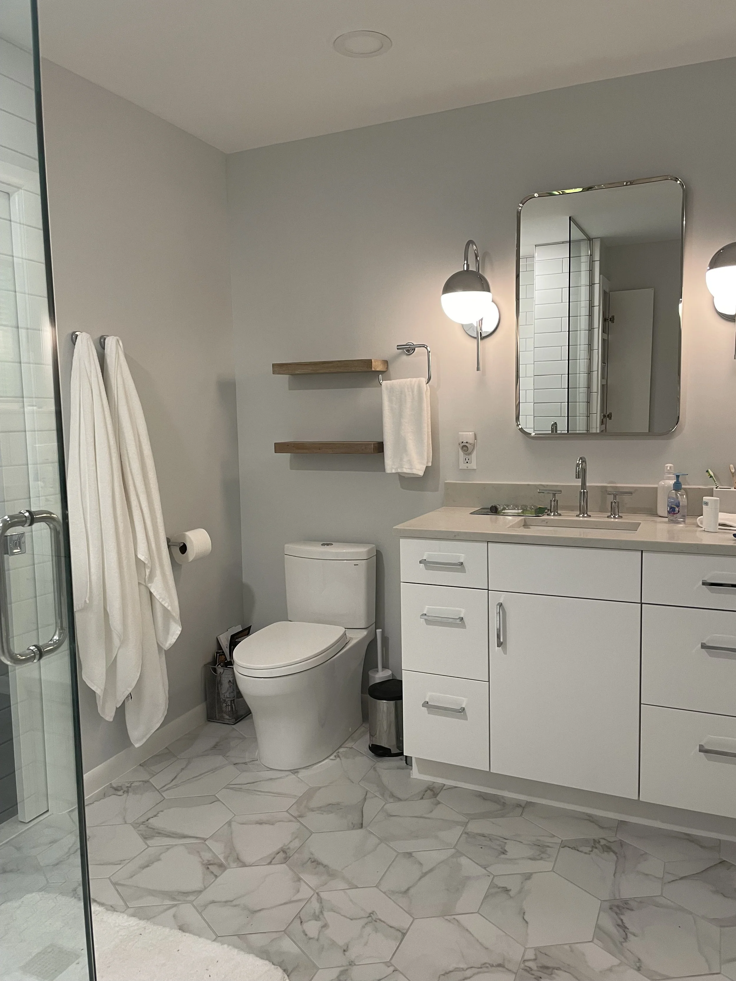 Modern bathroom with white vanity, marble hexagon floor tiles, wall-mounted mirror, wall lights, toilet, and a glass shower door.