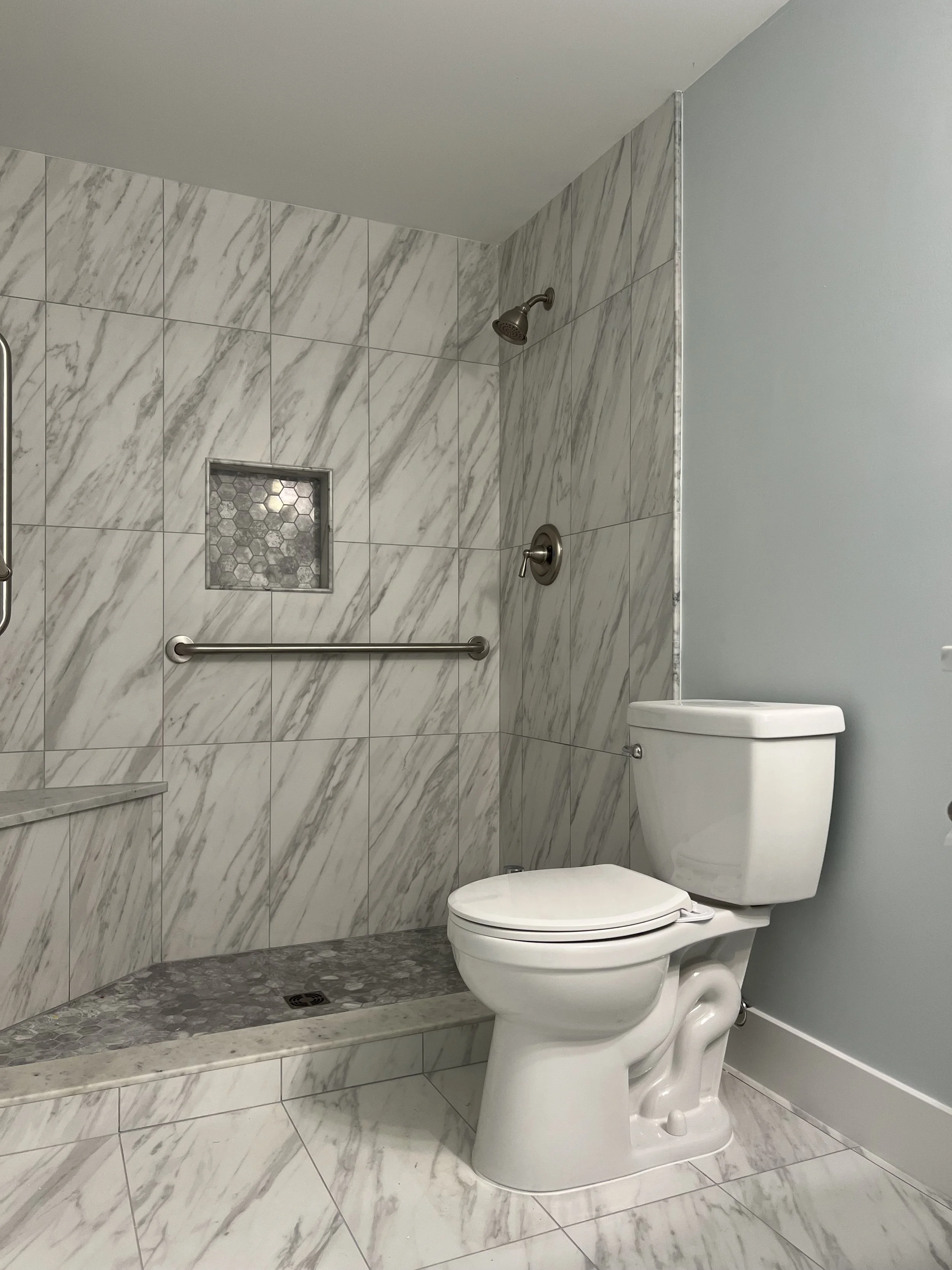 Bathroom with marble tiles, a toilet, a shower area with a grab bar, a small niche with hexagonal tiles, and a light blue wall.