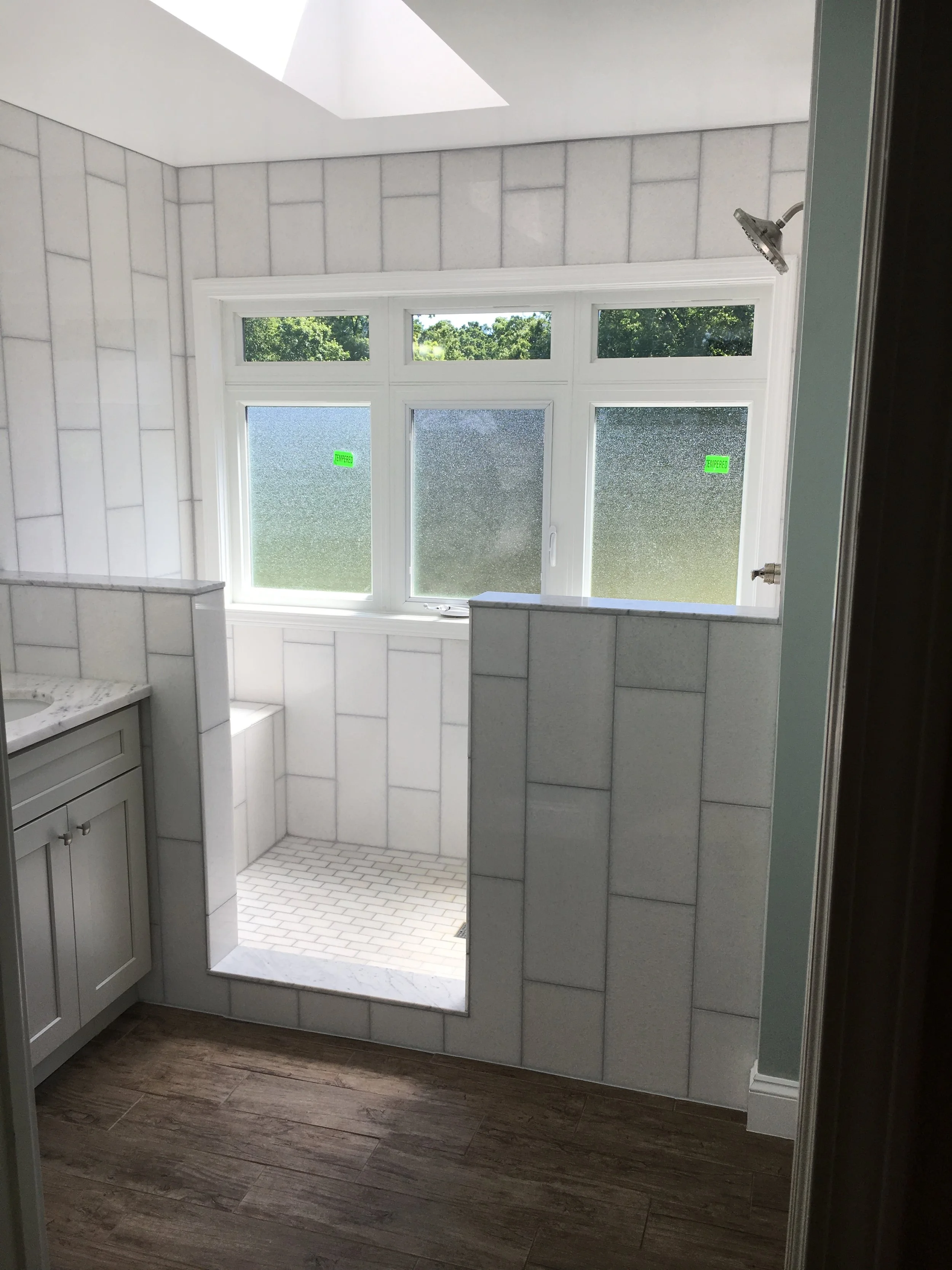 Bathroom with large frosted window, tiled walls, and a small shower area with a rain shower head. A vanity with a marble countertop is partially visible on the left. Wood-like flooring completes the space.